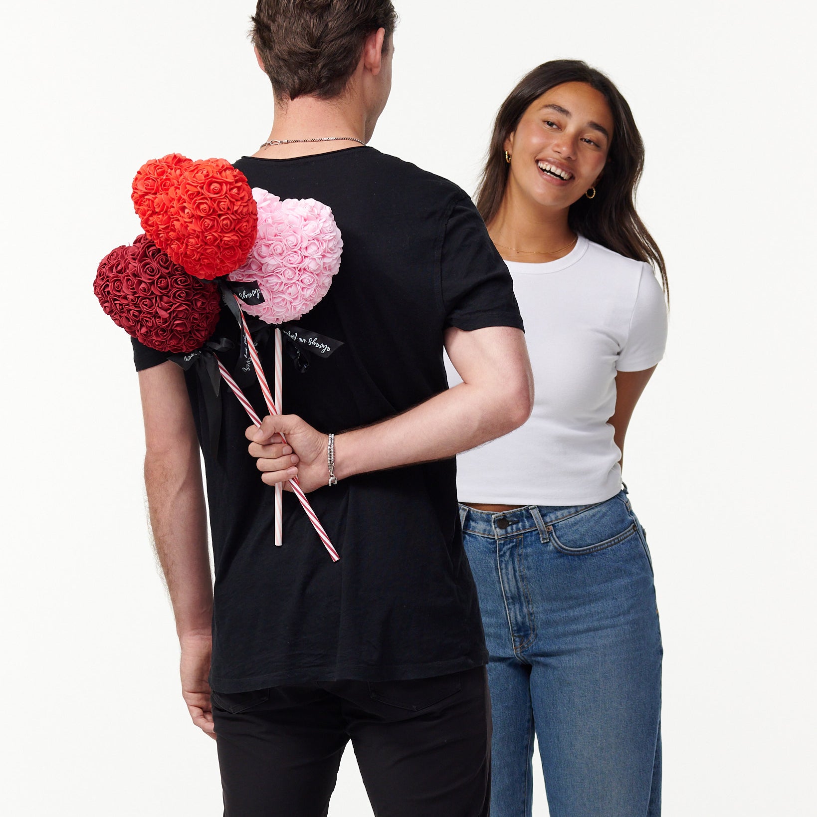 The image shows a male model in a black T-shirt, seen from behind, holding three artificial flower lollipops of red, pink, and burgundy behind his back, with a white female model in a white T-shirt and blue jeans smiling and facing him, seemingly unaware of the surprise. The lollipops have a striped stick with a bow, suggesting a playful or romantic gesture.