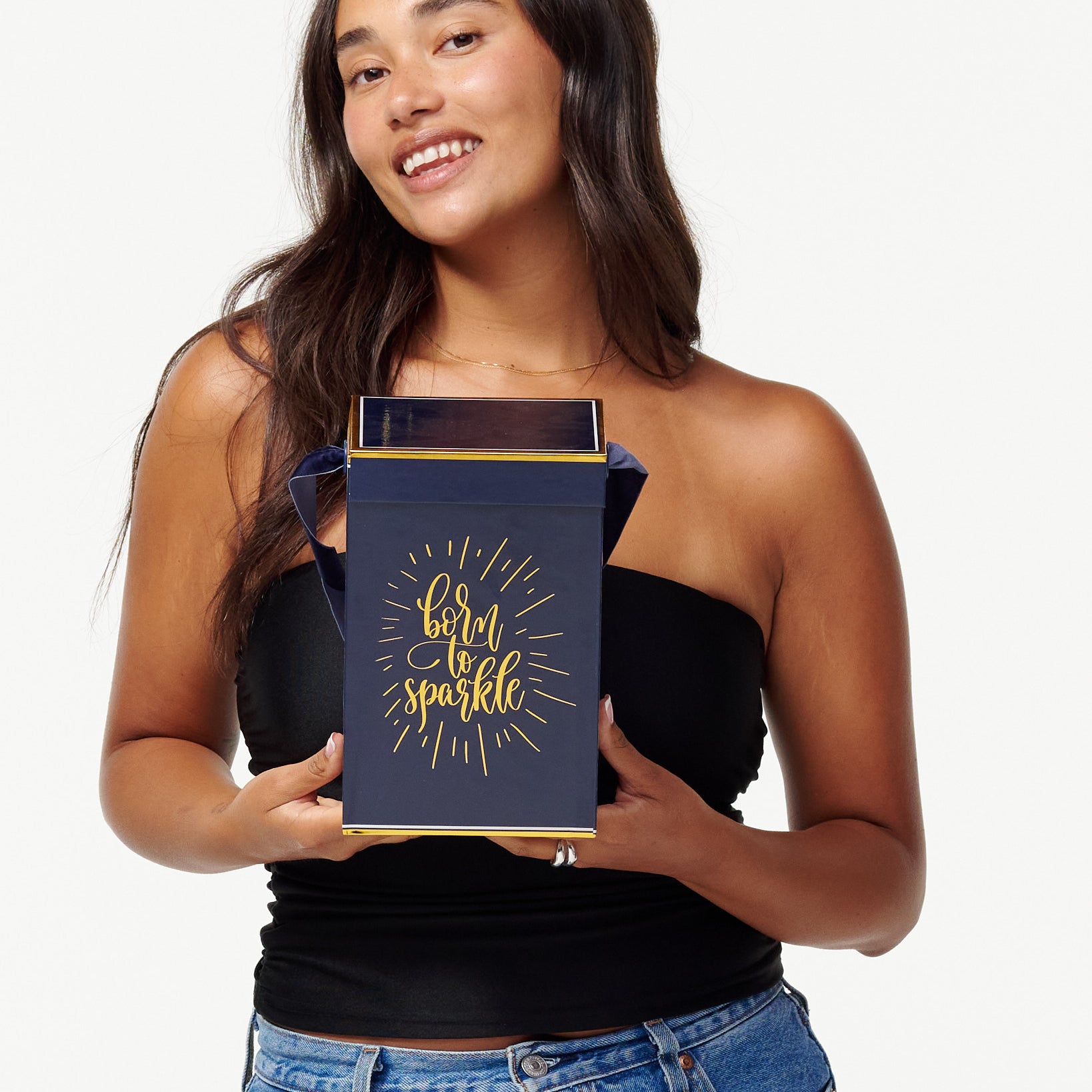 A smiling woman in a black top and jeans holds a blue box with the words "Born to Sparkle" in gold. She looks joyfully to the side, appearing content and playful.
