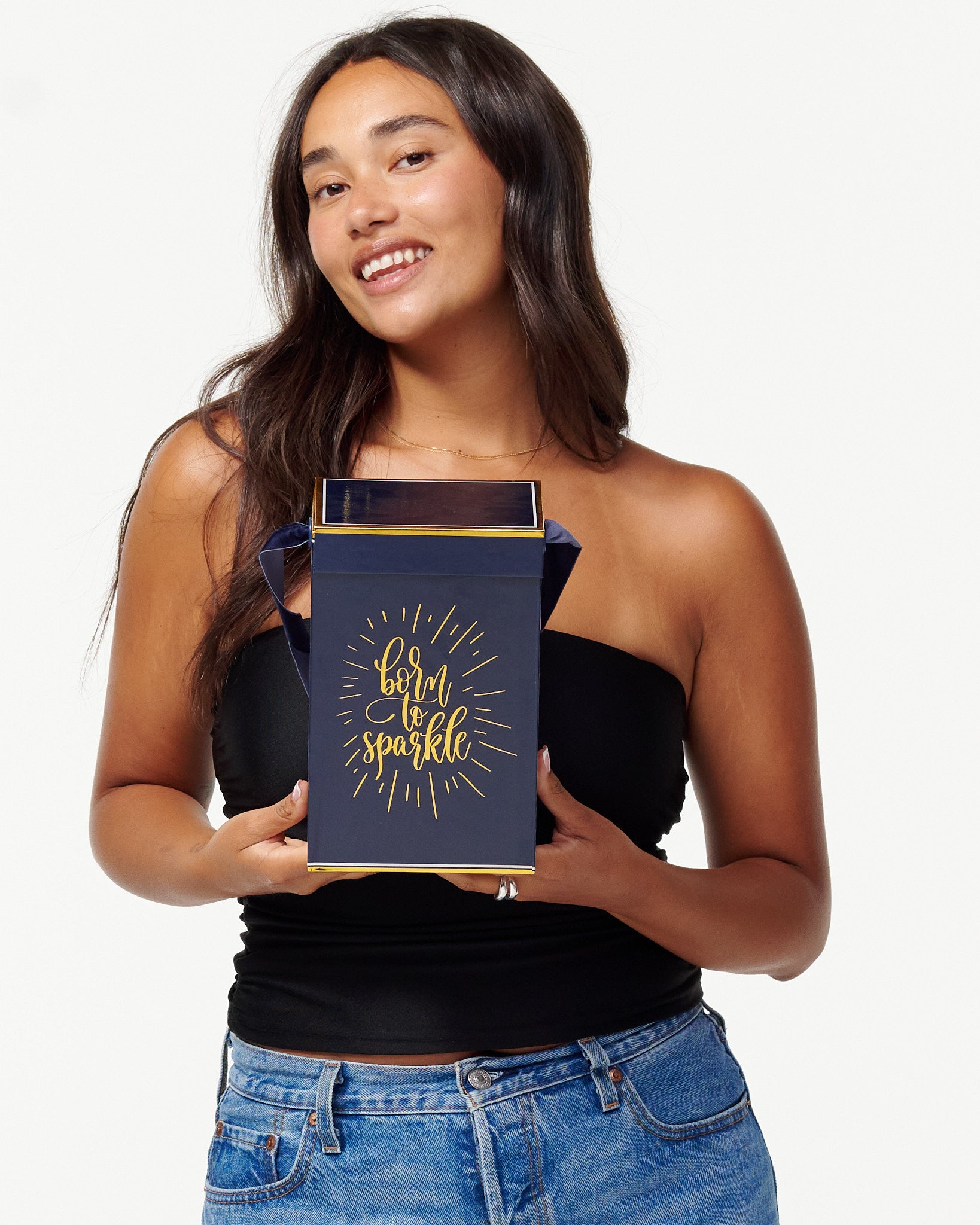 A smiling woman in a black top and jeans holds a blue box with the words "Born to Sparkle" in gold. She looks joyfully to the side, appearing content and playful.