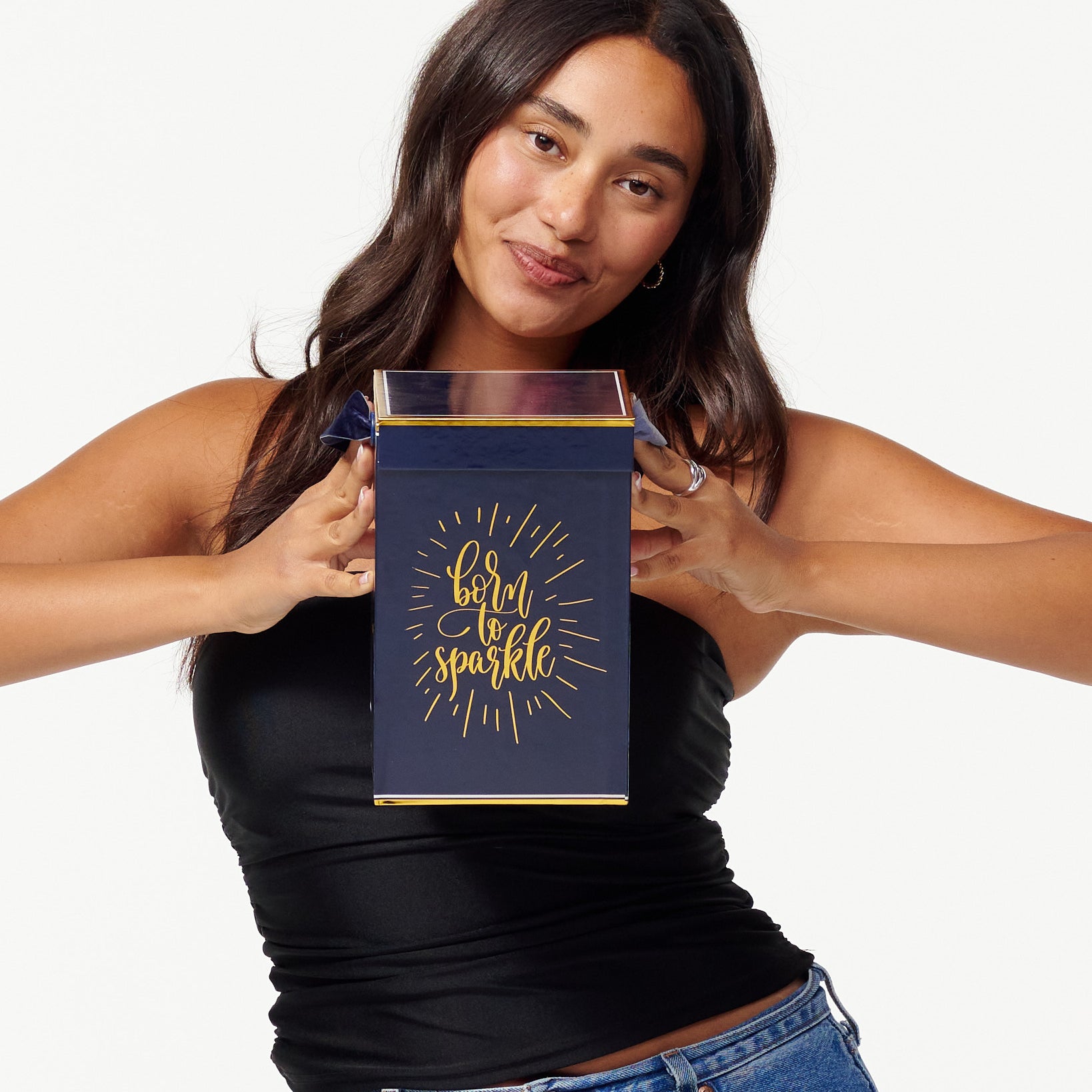 A smiling woman in a black top and jeans holds a blue box with the words "Born to Sparkle" in gold. She looks joyfully to the side, appearing content and playful.