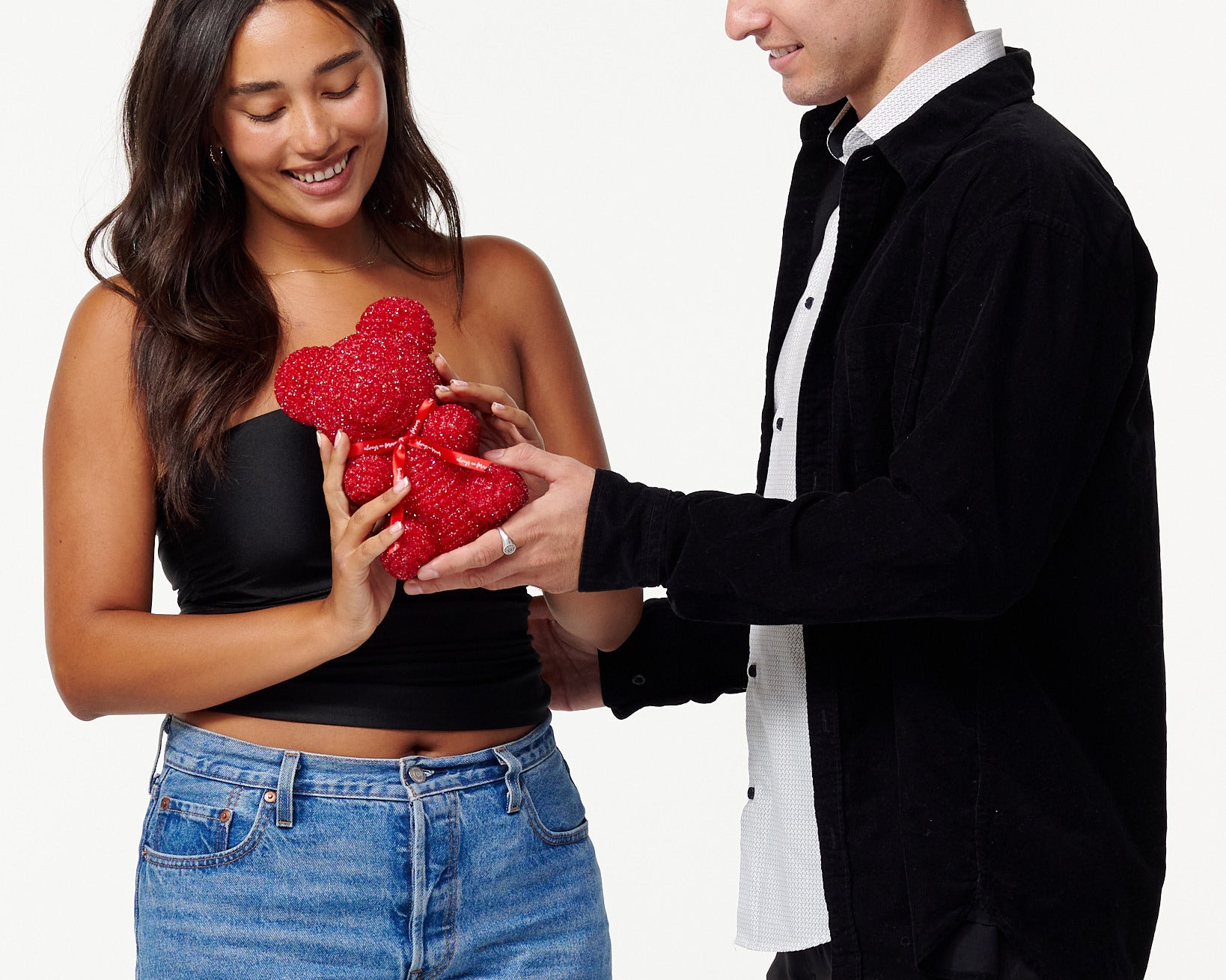 The image shows a woman and a man standing side by side. The woman is to the left, wearing a black cropped top and blue jeans, smiling down at a red, sparkly teddy bear she is holding. The man is to the right, dressed in a black jacket over a white shirt, also looking at the teddy bear with a smile, his hands gently touching the bear. Both individuals appear cheerful and are engaging with the teddy bear in a gentle manner.