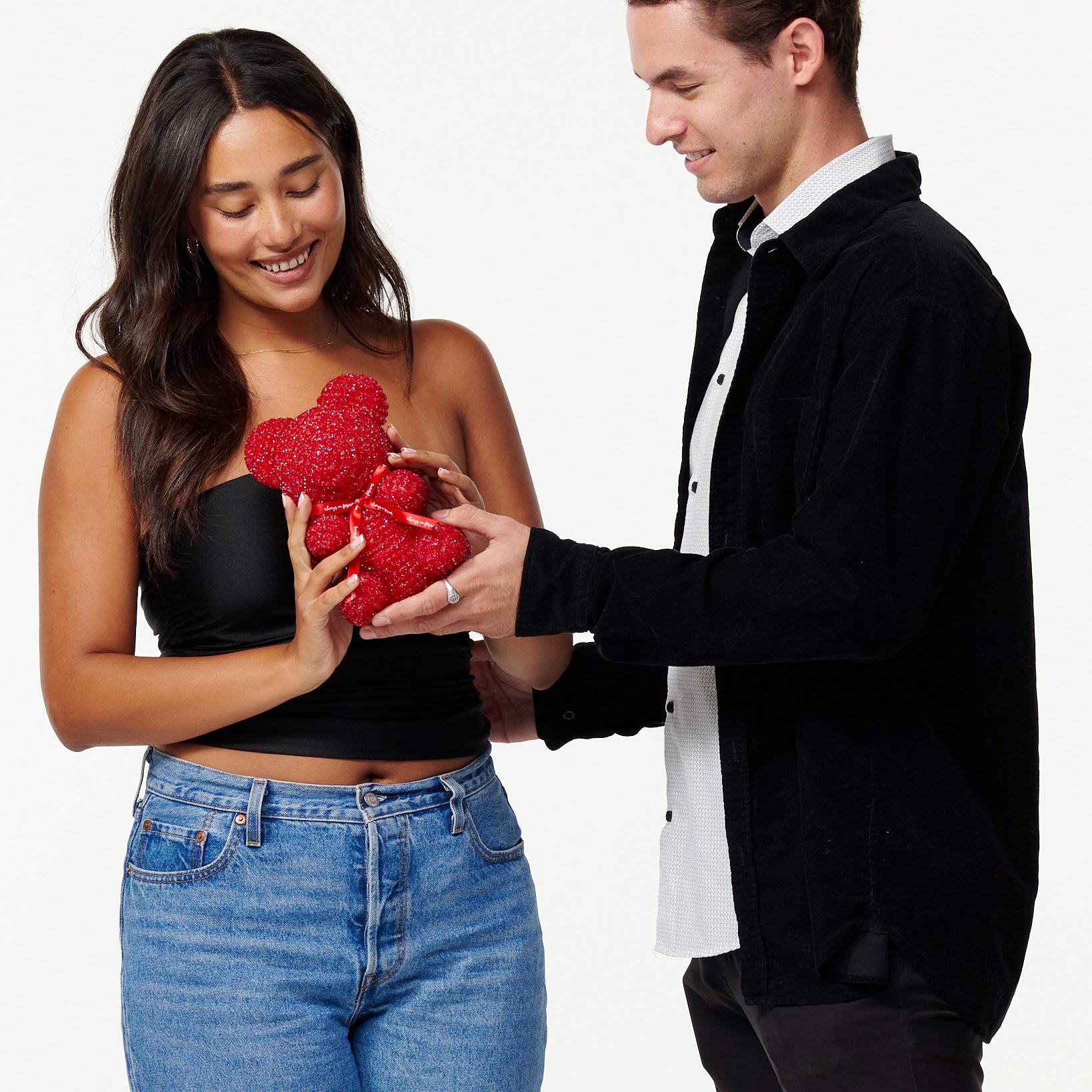 The image shows a woman and a man standing side by side. The woman is to the left, wearing a black cropped top and blue jeans, smiling down at a red, sparkly teddy bear she is holding. The man is to the right, dressed in a black jacket over a white shirt, also looking at the teddy bear with a smile, his hands gently touching the bear. Both individuals appear cheerful and are engaging with the teddy bear in a gentle manner.