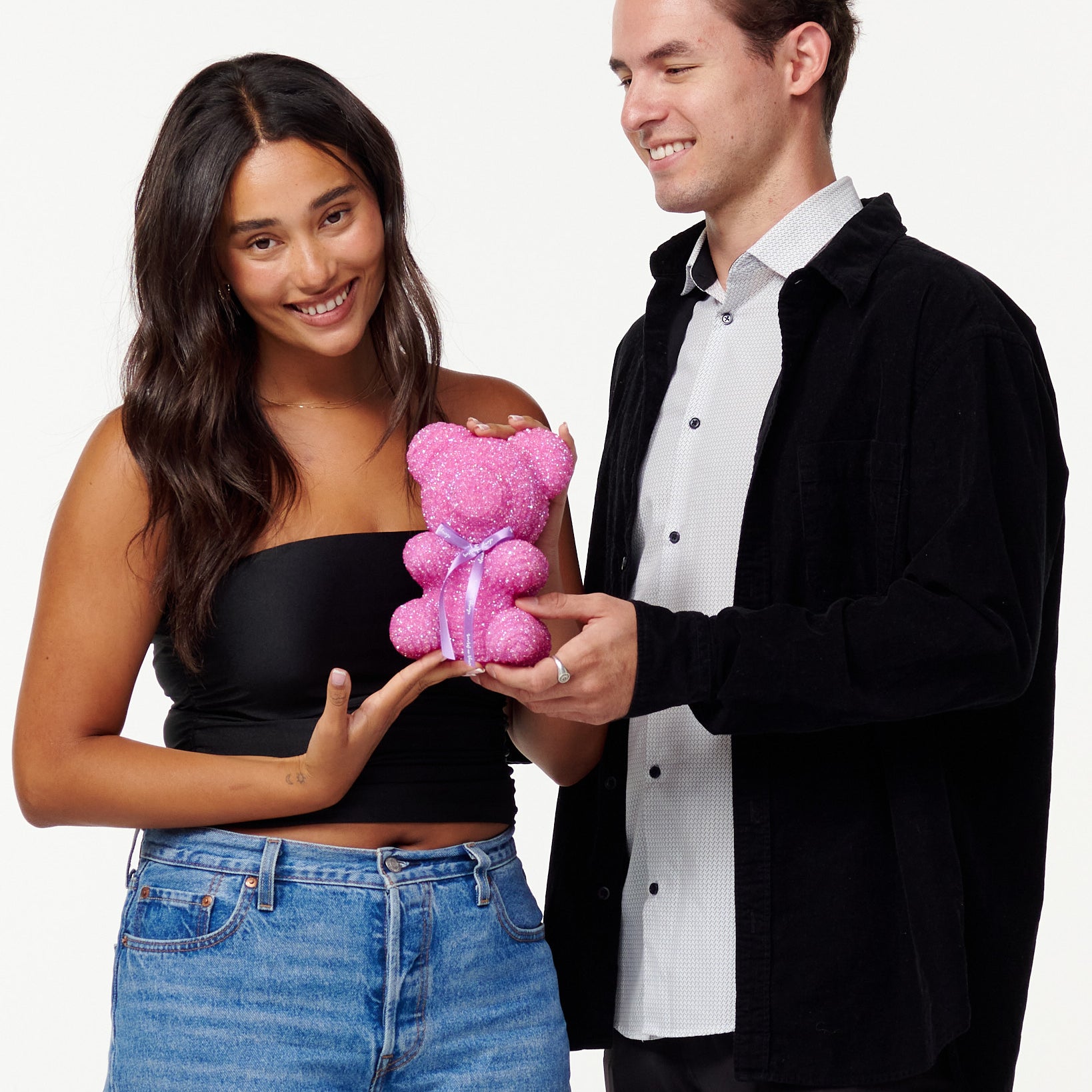 A woman in a black cropped top and blue jeans holds a pink, sparkly teddy bear with an admiring smile. Next to her, a man in a black jacket and white shirt looks at the teddy bear with a pleasant smile, his hand supporting the bear from beneath as they both enjoy the teddy bear's appearance.