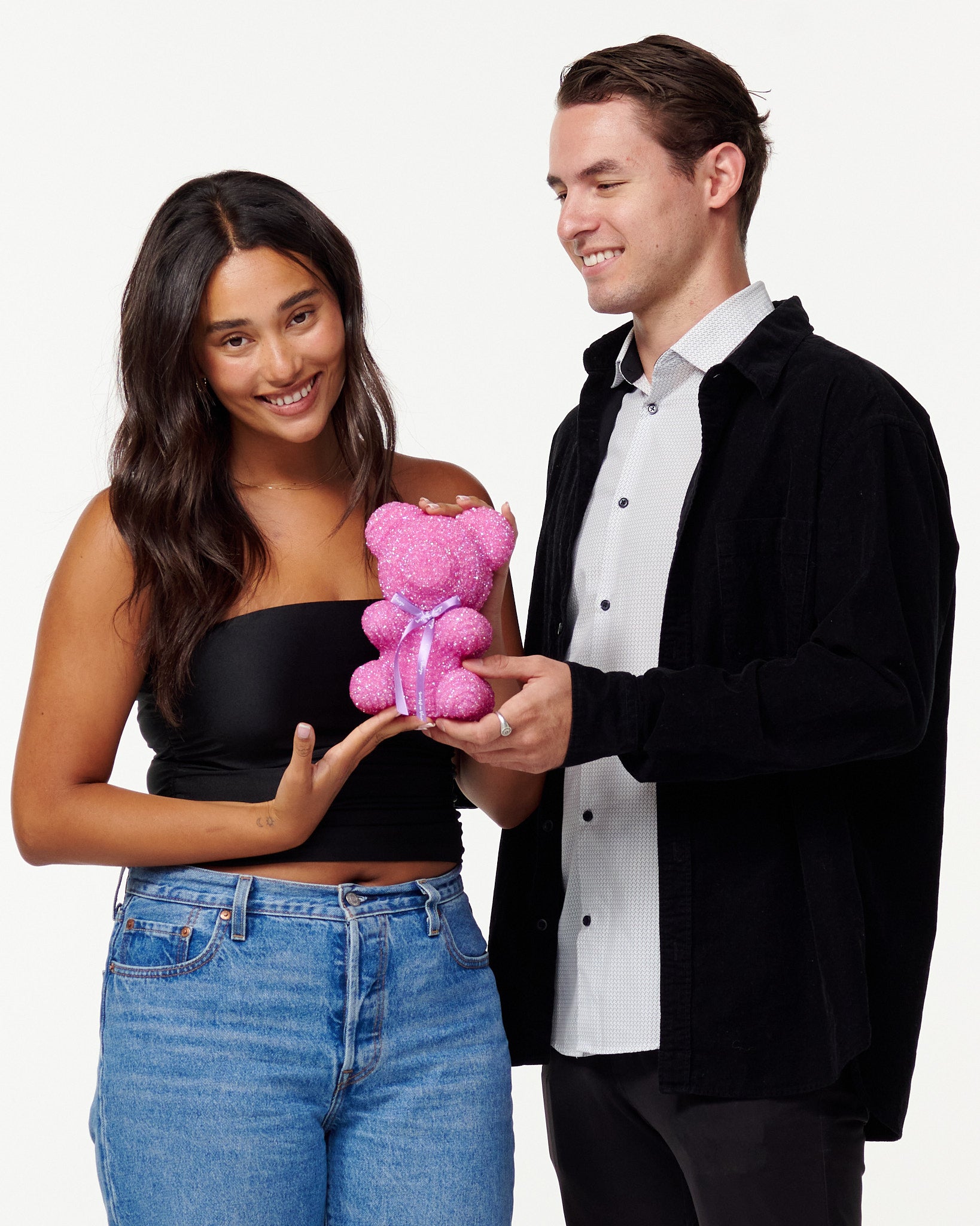 A woman in a black cropped top and blue jeans holds a pink, sparkly teddy bear with an admiring smile. Next to her, a man in a black jacket and white shirt looks at the teddy bear with a pleasant smile, his hand supporting the bear from beneath as they both enjoy the teddy bear's appearance.