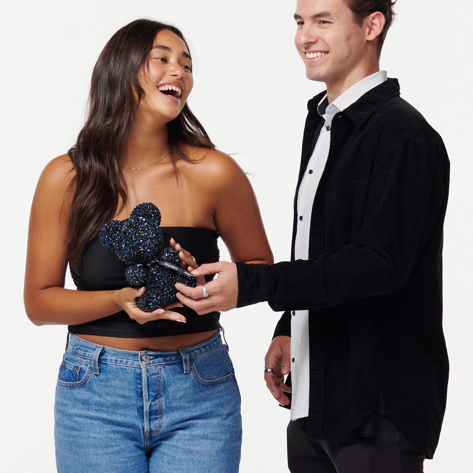 A woman in a black strapless top and jeans holds a glittery black flower bear, while a man in a black jacket over a white shirt smiles at her, touching the bear's paw. They both appear cheerful and are posing for the camera.