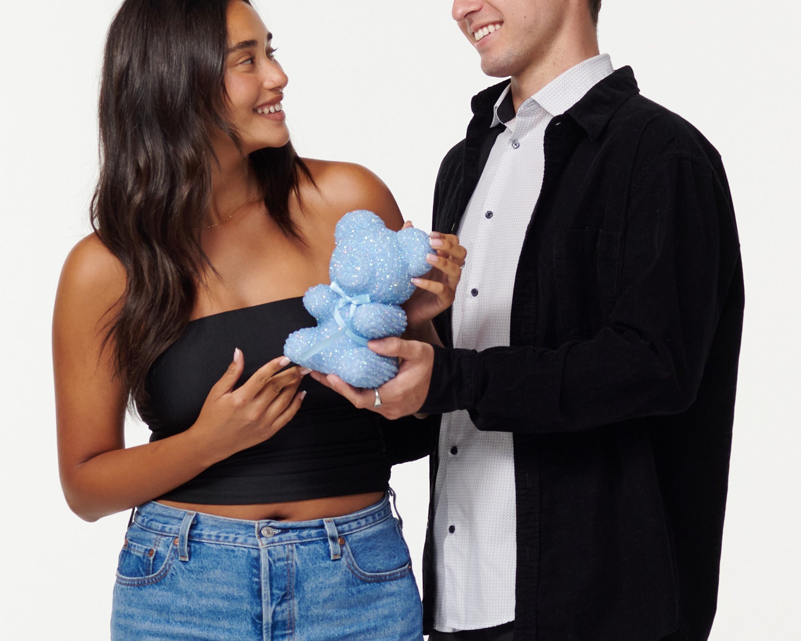 A woman in a black cropped top and blue jeans holds a blue, sparkly teddy bear, looking at it with a smile. Next to her, a man in a black jacket over a white shirt smiles as he looks at the teddy bear, his hands gently touching it. Both individuals appear engaged and happy with the teddy bear.