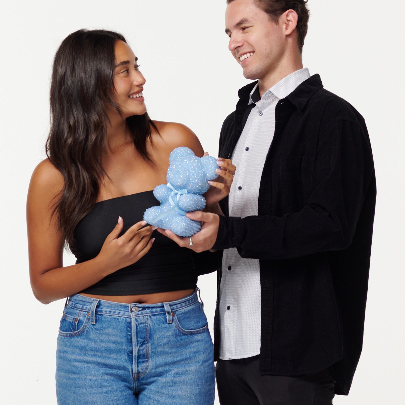 A woman in a black cropped top and blue jeans holds a blue, sparkly teddy bear, looking at it with a smile. Next to her, a man in a black jacket over a white shirt smiles as he looks at the teddy bear, his hands gently touching it. Both individuals appear engaged and happy with the teddy bear.