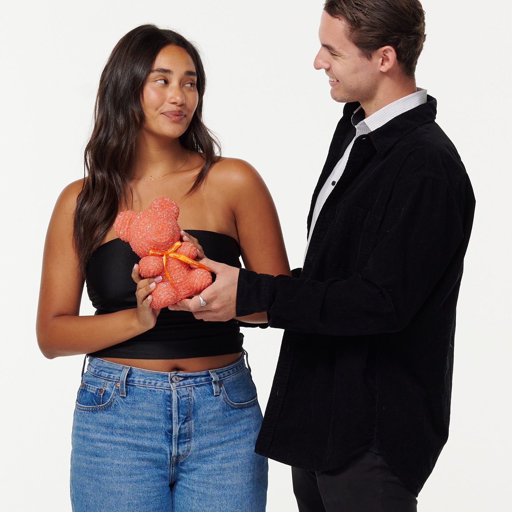A woman in a black cropped top and blue jeans is holding an pink, sparkly teddy bear and looking at it fondly. Beside her, a man in a black jacket and white shirt is also looking at the teddy bear with a smile, his hands placed on the bear as they both share a moment of joy.