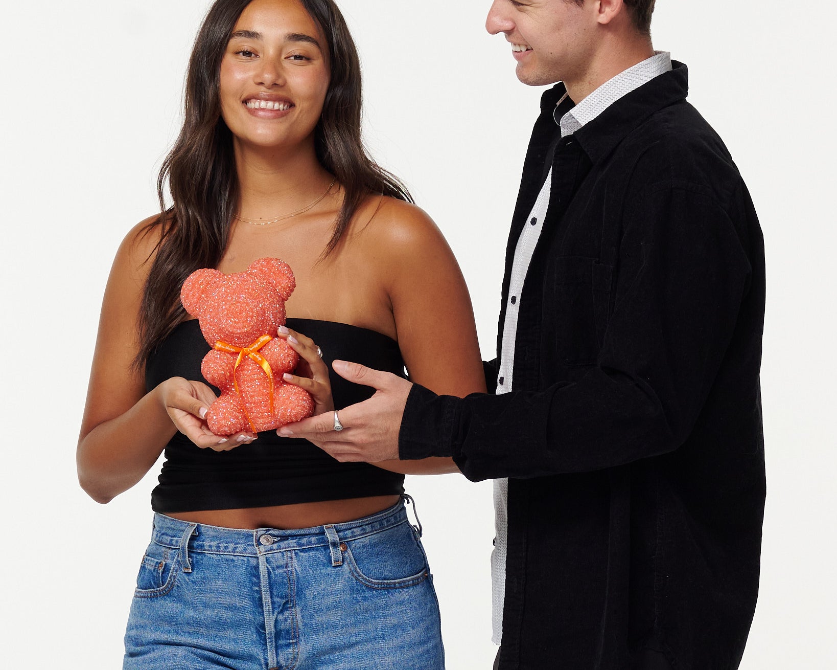 A woman in a black cropped top and blue jeans holds a blue, sparkly teddy bear, looking at it with a smile. Next to her, a man in a black jacket over a white shirt smiles as he looks at the teddy bear, his hands gently touching it. Both individuals appear engaged and happy with the teddy bear.