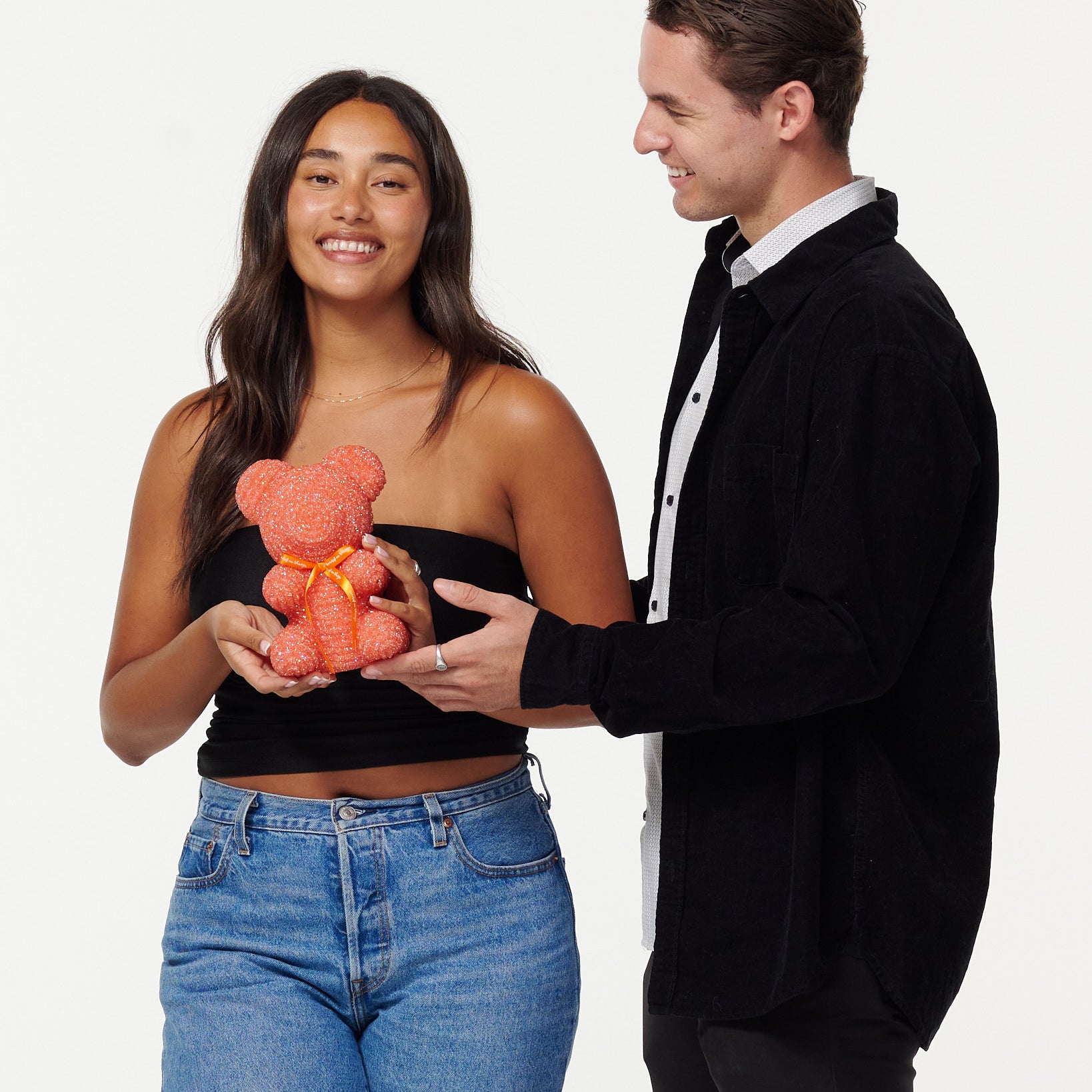 A woman in a black cropped top and blue jeans holds a blue, sparkly teddy bear, looking at it with a smile. Next to her, a man in a black jacket over a white shirt smiles as he looks at the teddy bear, his hands gently touching it. Both individuals appear engaged and happy with the teddy bear.