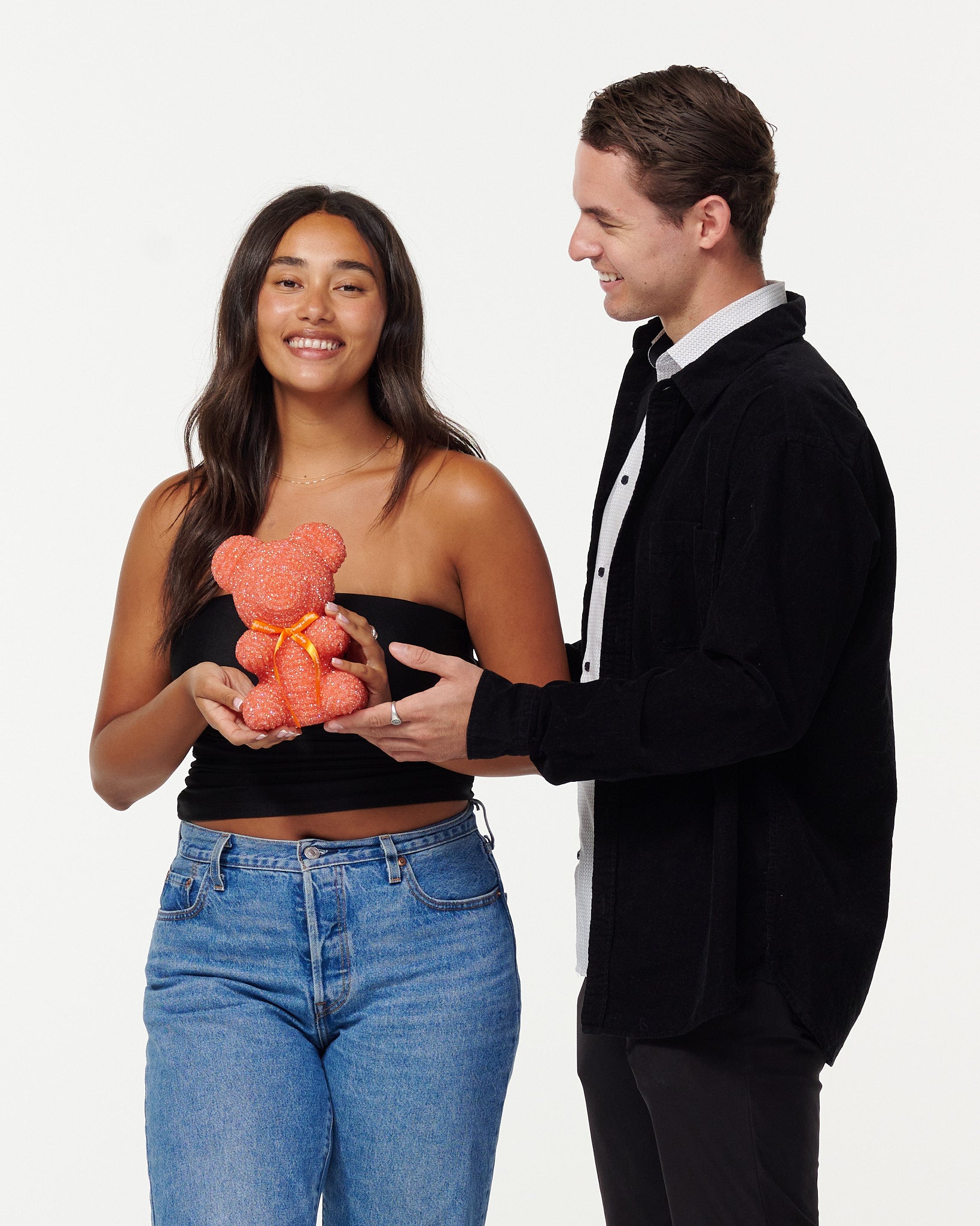 A woman in a black cropped top and blue jeans holds a blue, sparkly teddy bear, looking at it with a smile. Next to her, a man in a black jacket over a white shirt smiles as he looks at the teddy bear, his hands gently touching it. Both individuals appear engaged and happy with the teddy bear.