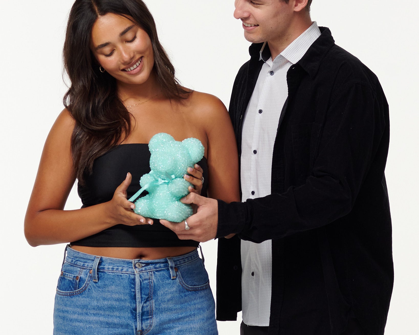 A woman in a black top and jeans admires a teal glitter bear, while a smiling man in a black jacket and white shirt gently touches the bear's arm. They appear to be in a light-hearted, affectionate interaction.
