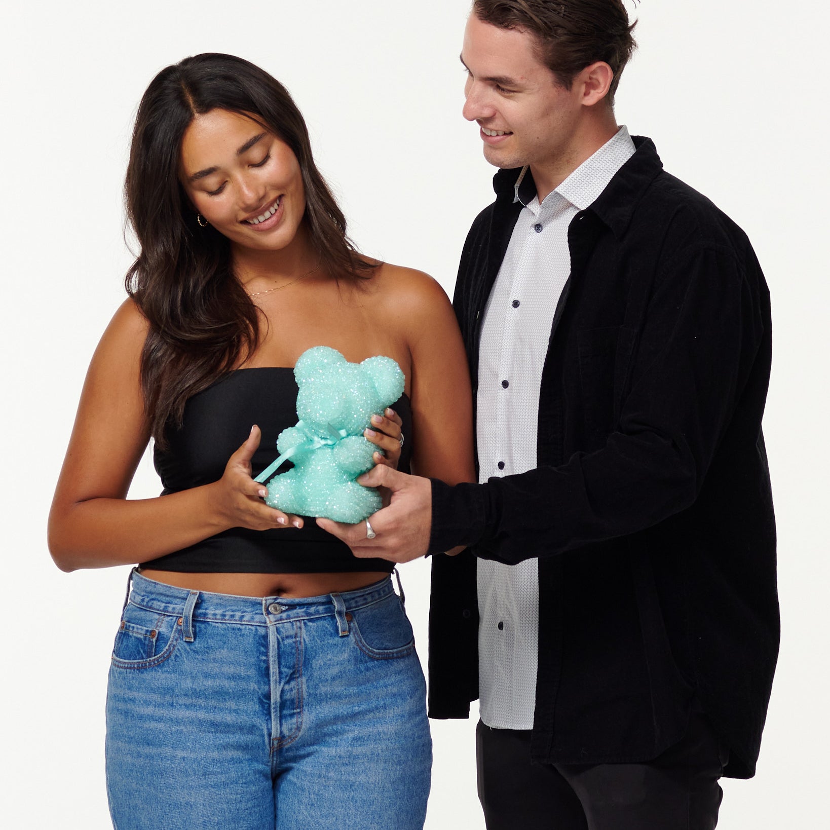 A woman in a black top and jeans admires a teal glitter bear, while a smiling man in a black jacket and white shirt gently touches the bear's arm. They appear to be in a light-hearted, affectionate interaction.