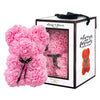 Pink, rose-shaped faux bloom bear with a black ribbon around its neck that includes the text "Just for You". The bear is placed next to a stylish white gift box with black accents, which has a clear window. The box comes with a black carrying handle. This intricately crafted floral bear serves as a perfect gift for various occasions such as anniversaries, birthdays, graduations, Christmas, Valentine's Day, or simply as a beautiful decorative item for those special moments.