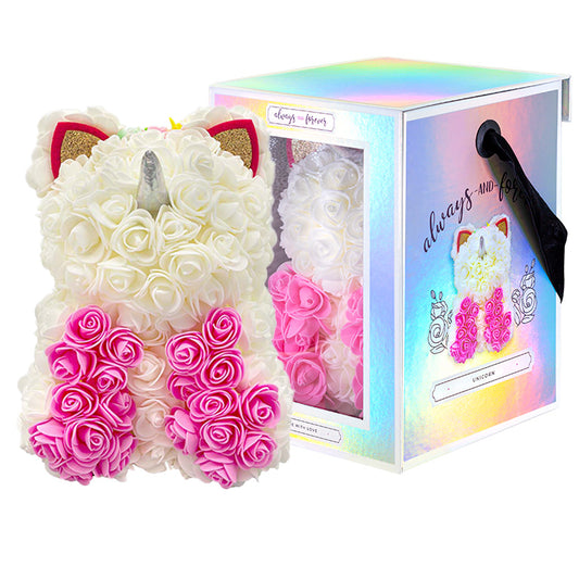 An artificial flower unicorn featuring white roses with pink roses for its mane, golden ears, and a silver horn. It's beside its holographic box packaging that has a "always and forever" inscription and a transparent window revealing another unicorn, labeled "UNICORN" with a "Made with Love" tag at the bottom.