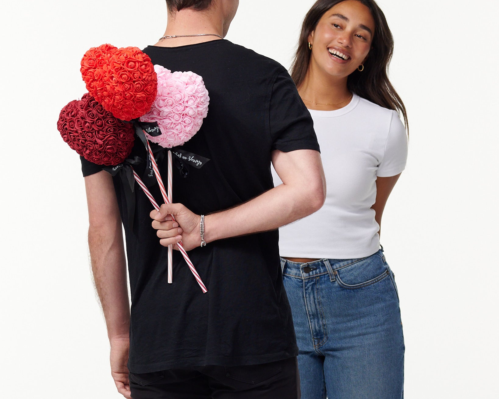 The image shows a male model in a black T-shirt, seen from behind, holding three artificial flower lollipops of red, pink, and burgundy behind his back, with a white female model in a white T-shirt and blue jeans smiling and facing him, seemingly unaware of the surprise. The lollipops have a striped stick with a bow, suggesting a playful or romantic gesture.