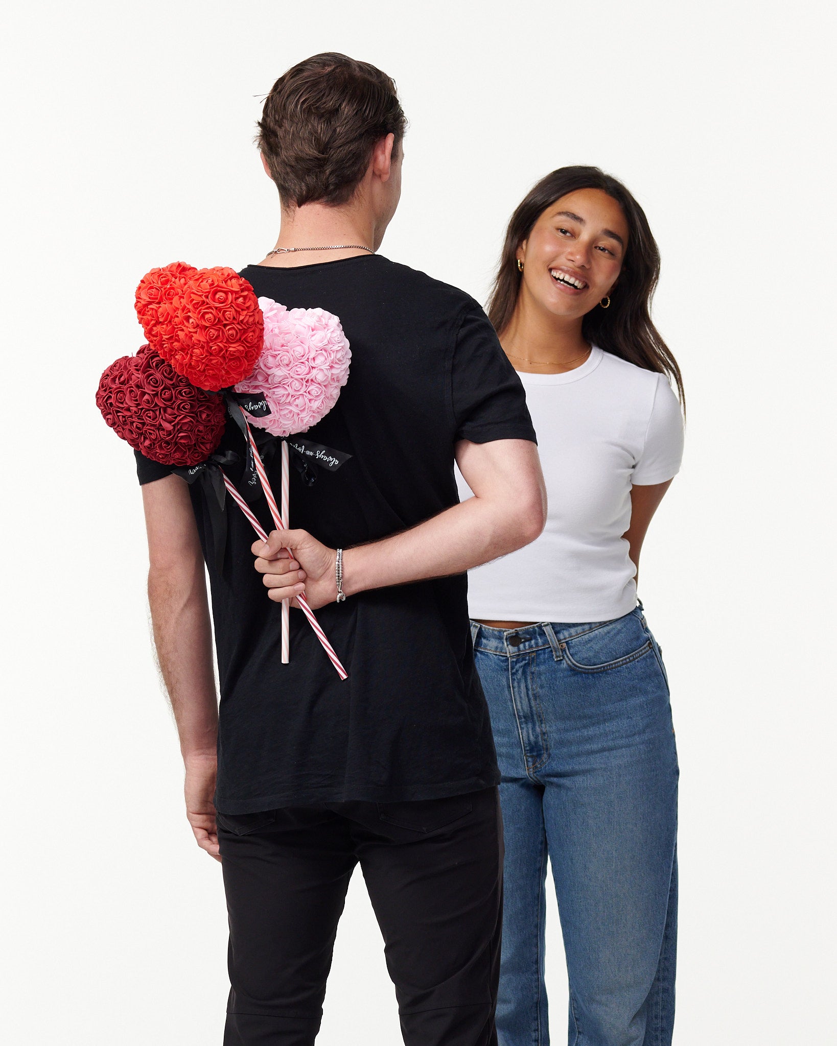 The image shows a male model in a black T-shirt, seen from behind, holding three artificial flower lollipops of red, pink, and burgundy behind his back, with a white female model in a white T-shirt and blue jeans smiling and facing him, seemingly unaware of the surprise. The lollipops have a striped stick with a bow, suggesting a playful or romantic gesture.