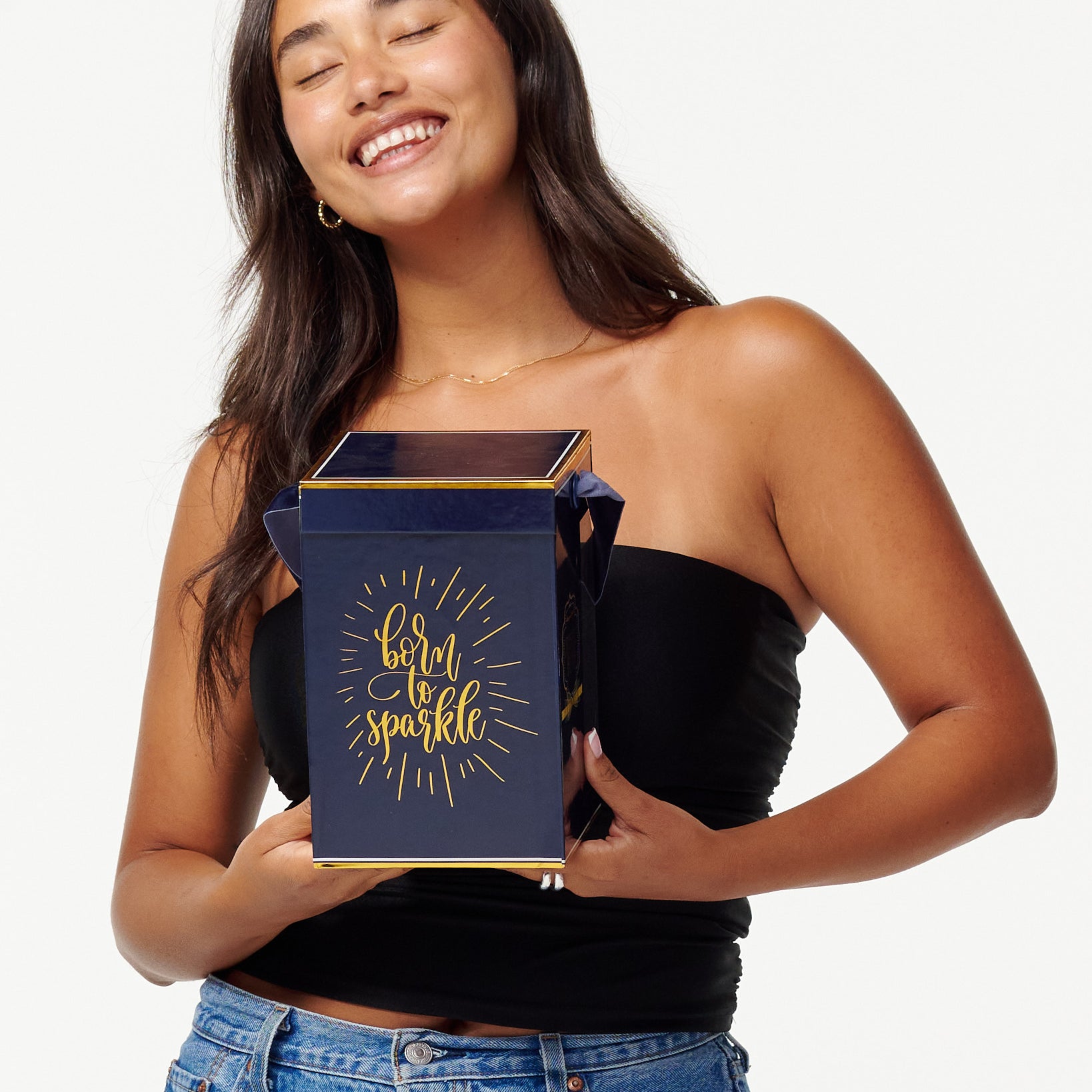 A smiling woman in a black top and jeans holds a blue box with the words "Born to Sparkle" in gold. She looks joyfully to the side, appearing content and playful.