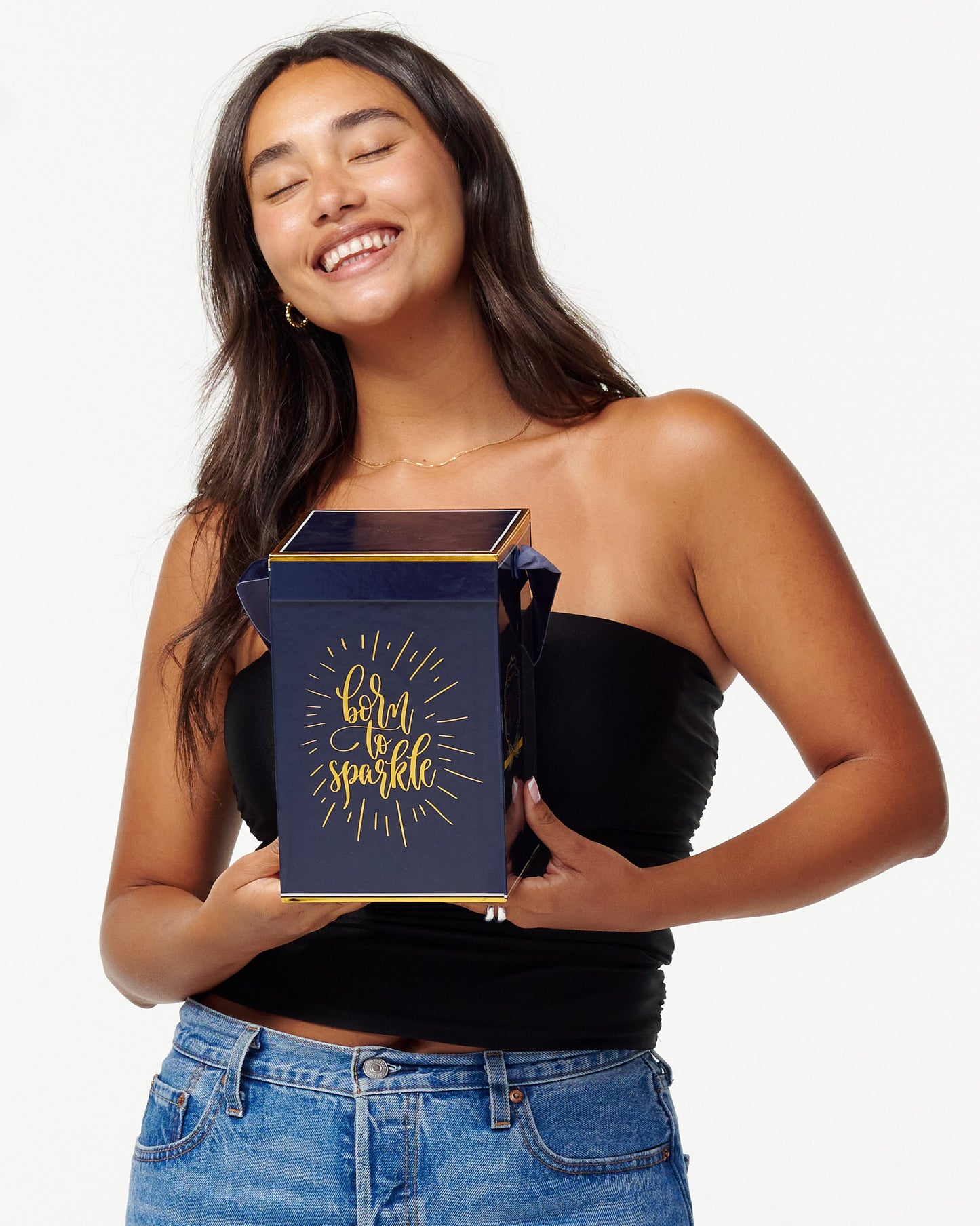 A smiling woman in a black top and jeans holds a blue box with the words "Born to Sparkle" in gold. She looks joyfully to the side, appearing content and playful.