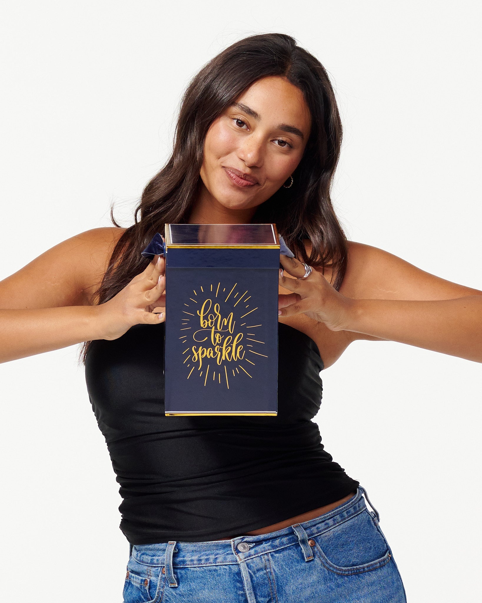 A smiling woman in a black top and jeans holds a blue box with the words "Born to Sparkle" in gold. She looks joyfully to the side, appearing content and playful.