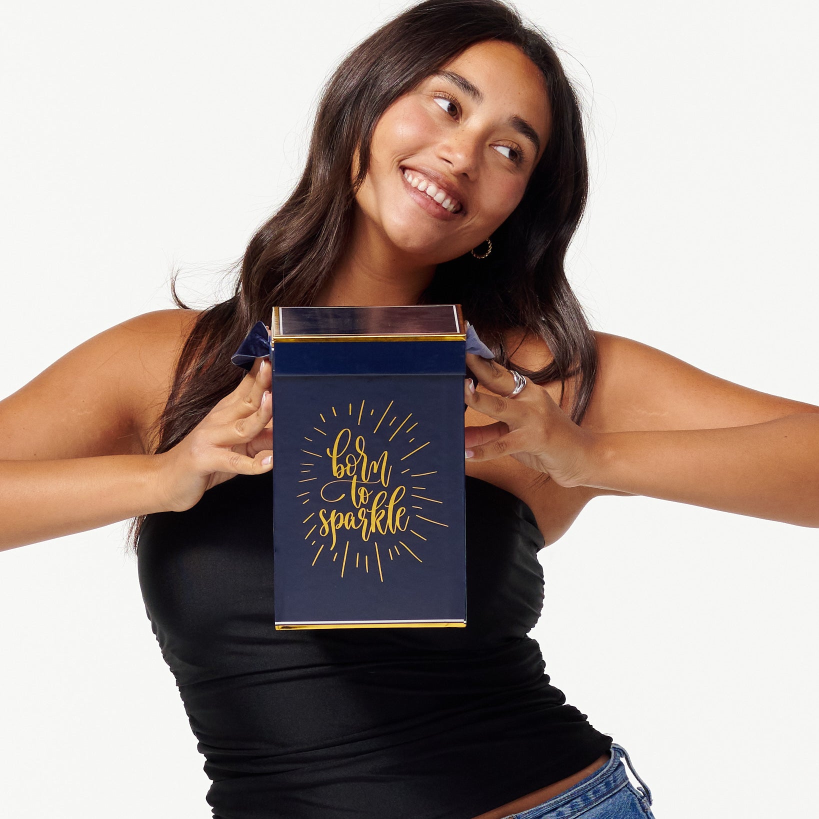 A smiling woman in a black top and jeans holds a blue box with the words "Born to Sparkle" in gold. She looks joyfully to the side, appearing content and playful.