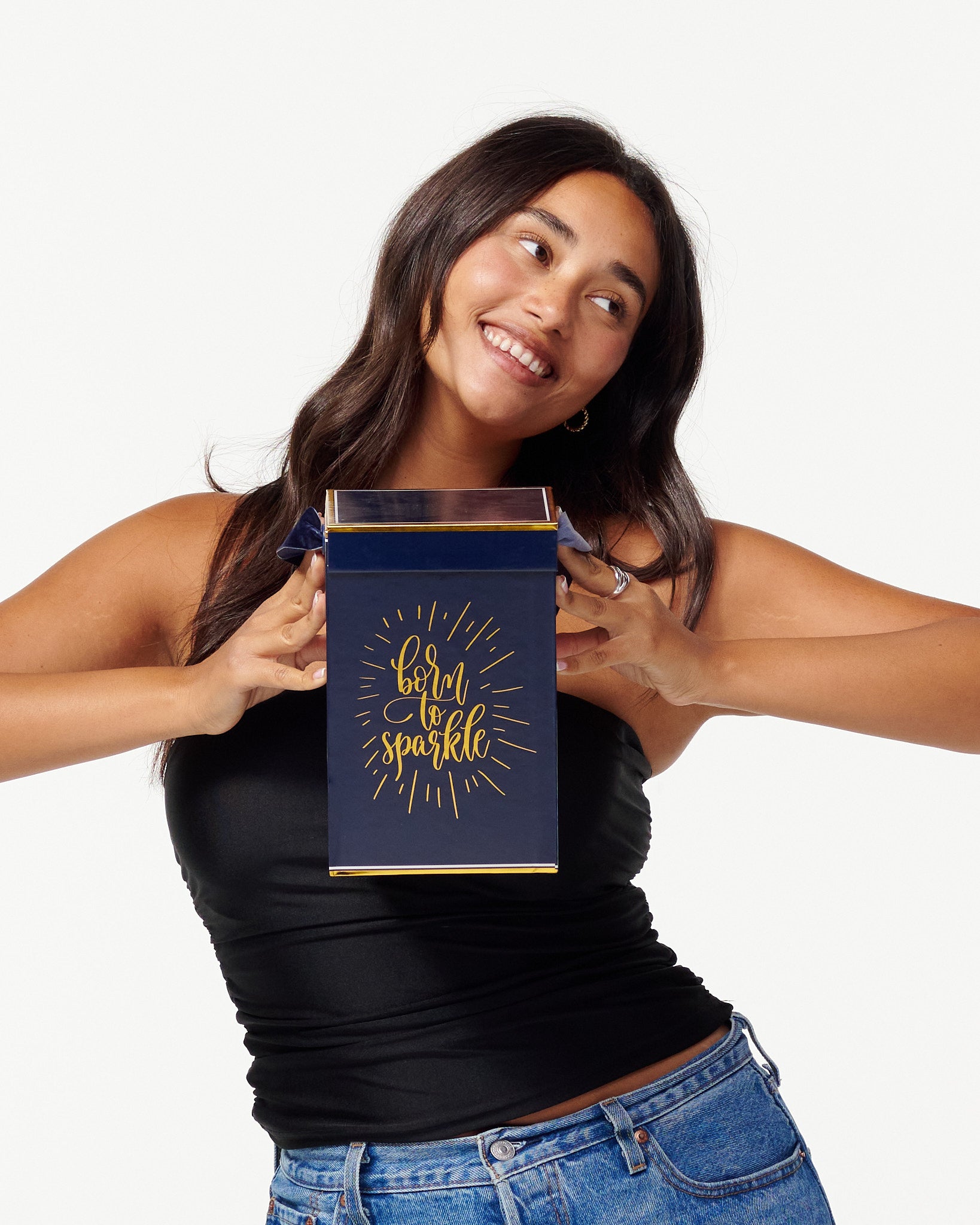 A smiling woman in a black top and jeans holds a blue box with the words "Born to Sparkle" in gold. She looks joyfully to the side, appearing content and playful.