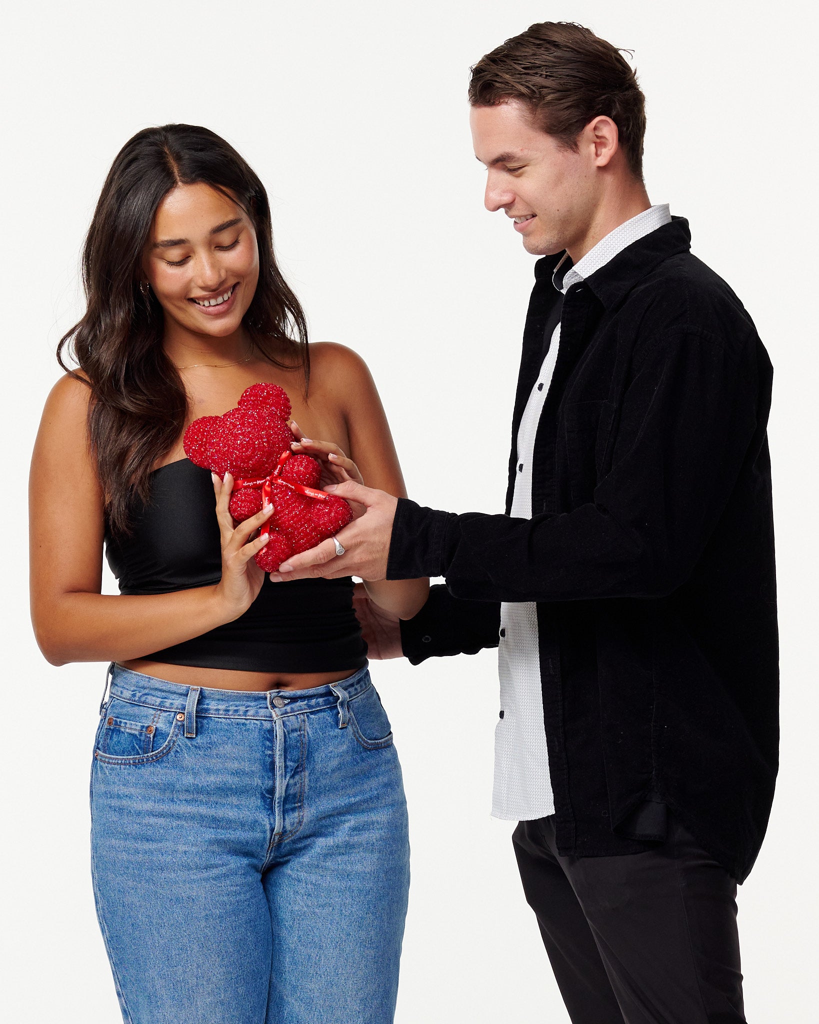  The image shows a woman and a man standing side by side. The woman is to the left, wearing a black cropped top and blue jeans, smiling down at a red, sparkly teddy bear she is holding. The man is to the right, dressed in a black jacket over a white shirt, also looking at the teddy bear with a smile, his hands gently touching the bear. Both individuals appear cheerful and are engaging with the teddy bear in a gentle manner. 