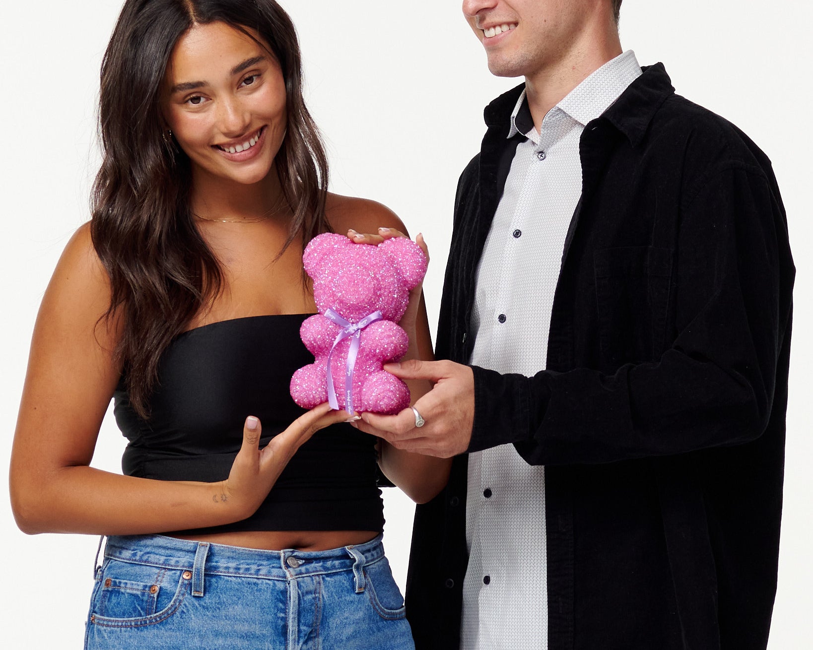A woman in a black cropped top and blue jeans holds a pink, sparkly teddy bear with an admiring smile. Next to her, a man in a black jacket and white shirt looks at the teddy bear with a pleasant smile, his hand supporting the bear from beneath as they both enjoy the teddy bear's appearance.