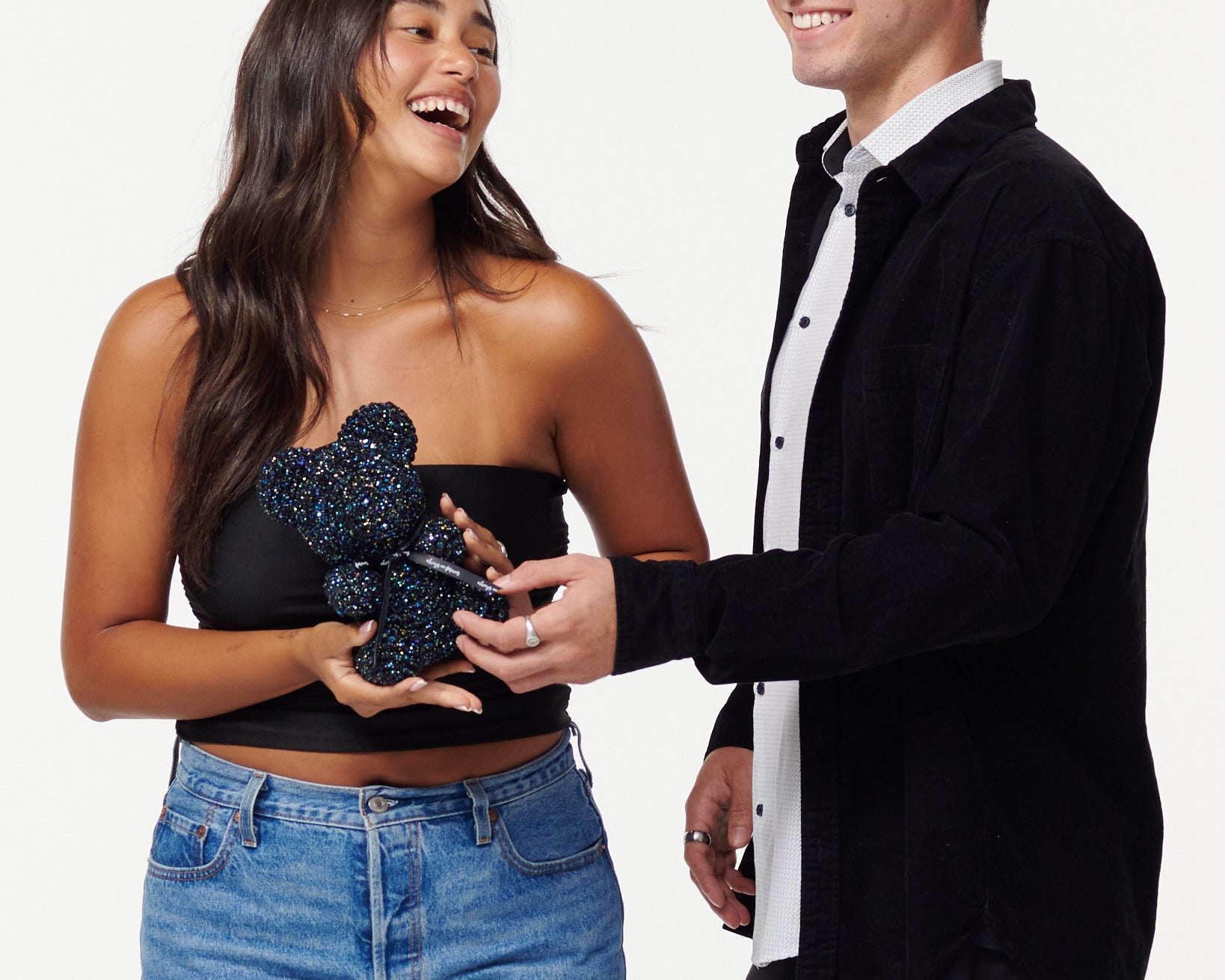 A woman in a black strapless top and jeans holds a glittery black flower bear, while a man in a black jacket over a white shirt smiles at her, touching the bear's paw. They both appear cheerful and are posing for the camera.