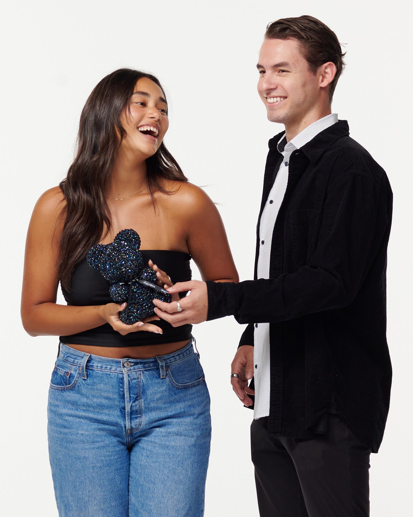 A woman in a black strapless top and jeans holds a glittery black flower bear, while a man in a black jacket over a white shirt smiles at her, touching the bear's paw. They both appear cheerful and are posing for the camera.