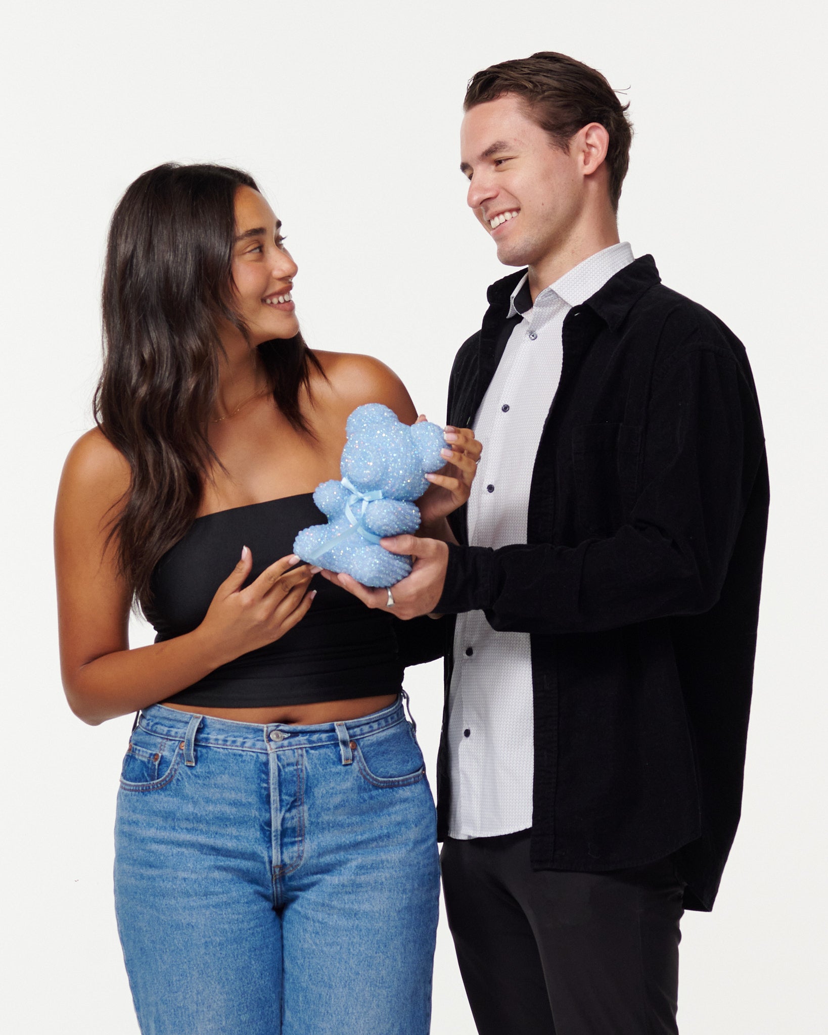 A woman in a black cropped top and blue jeans holds a blue, sparkly teddy bear, looking at it with a smile. Next to her, a man in a black jacket over a white shirt smiles as he looks at the teddy bear, his hands gently touching it. Both individuals appear engaged and happy with the teddy bear.