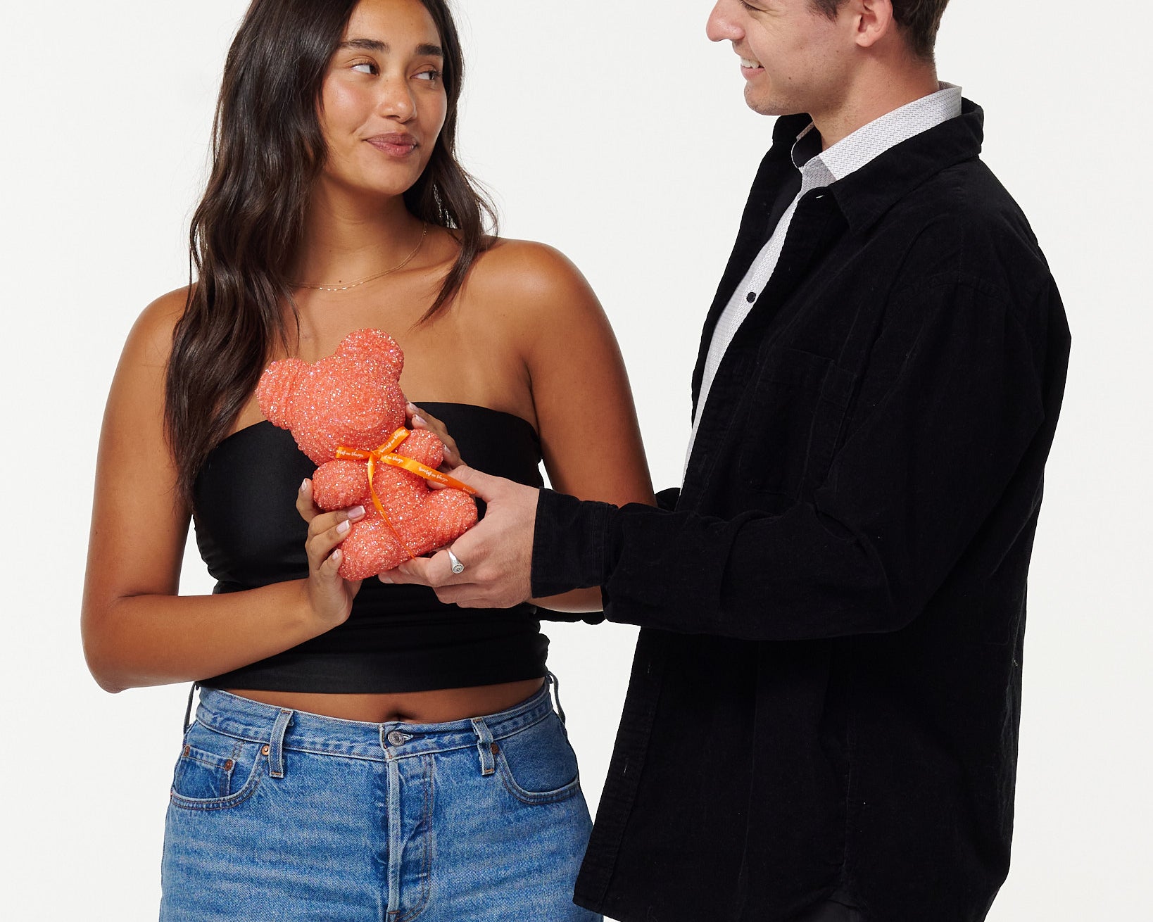 A woman in a black cropped top and blue jeans is holding an pink, sparkly teddy bear and looking at it fondly. Beside her, a man in a black jacket and white shirt is also looking at the teddy bear with a smile, his hands placed on the bear as they both share a moment of joy.