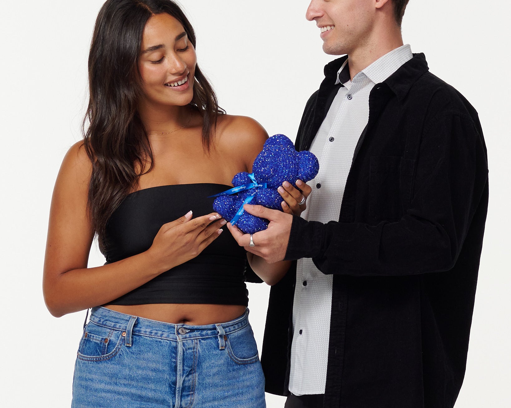 A woman in a black cropped top and blue jeans admires a blue, sparkly teddy bear with a bright blue ribbon, smiling at it. A man in a black jacket and white shirt stands beside her, looking at the teddy bear and smiling, his hands placed on the bear, as they share a moment of appreciation.
