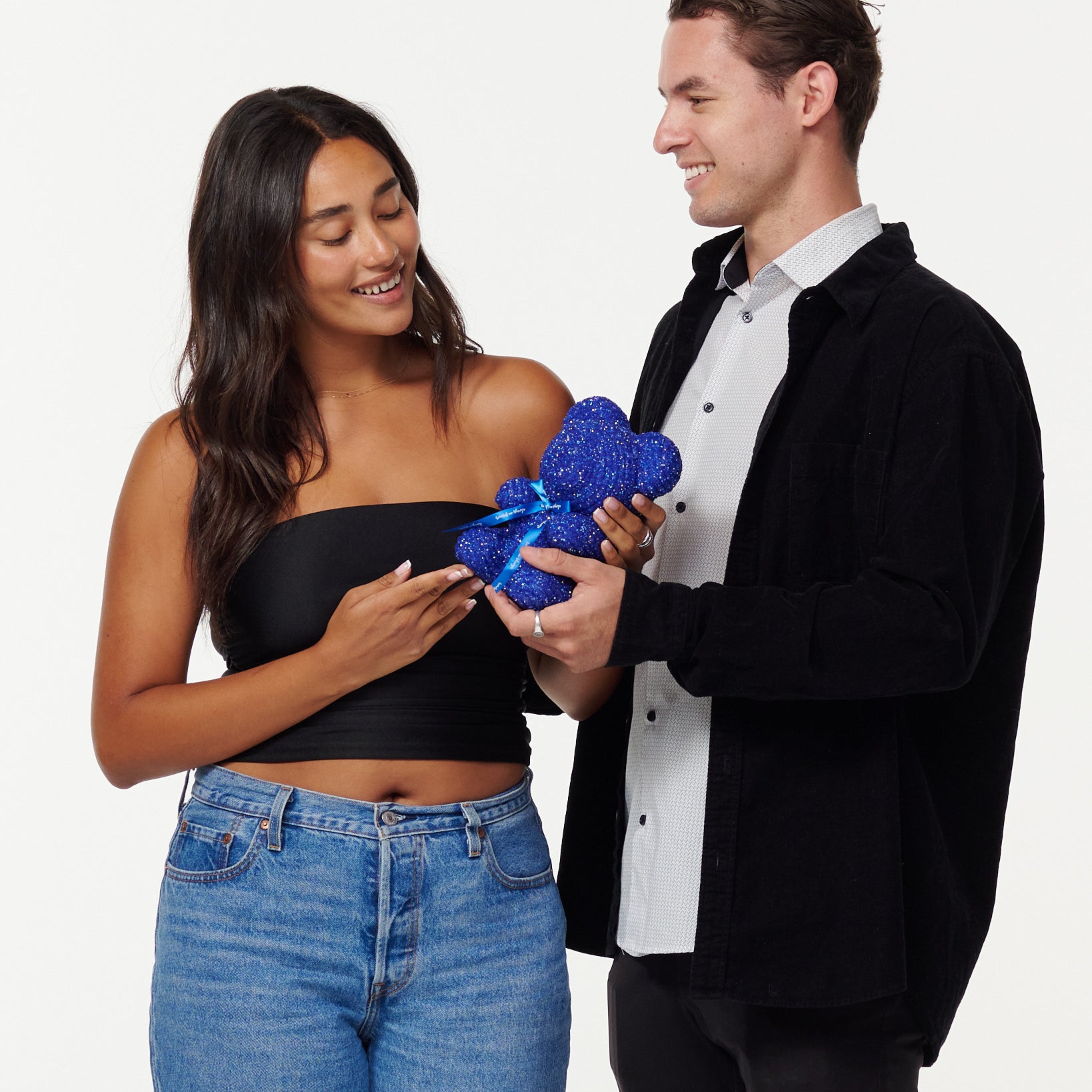 A woman in a black cropped top and blue jeans admires a blue, sparkly teddy bear with a bright blue ribbon, smiling at it. A man in a black jacket and white shirt stands beside her, looking at the teddy bear and smiling, his hands placed on the bear, as they share a moment of appreciation.