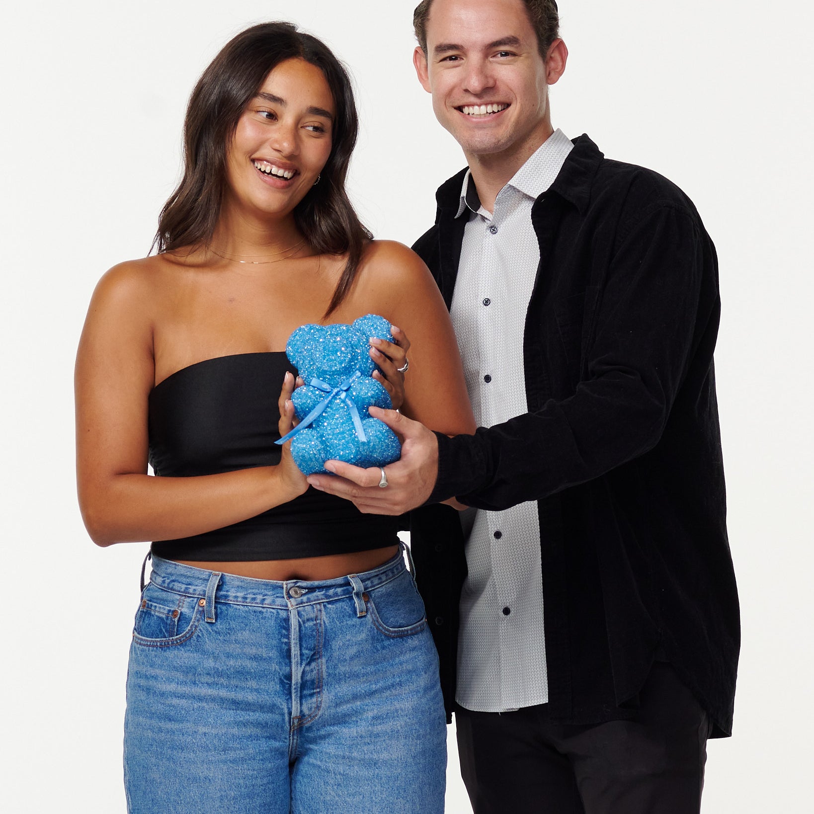 A woman in a black cropped top and blue jeans cradles a blue, sparkly teddy bear with a gentle smile. Next to her, a man in a black jacket and white shirt shares a warm smile, his hands on the teddy bear, as they both enjoy the moment together.