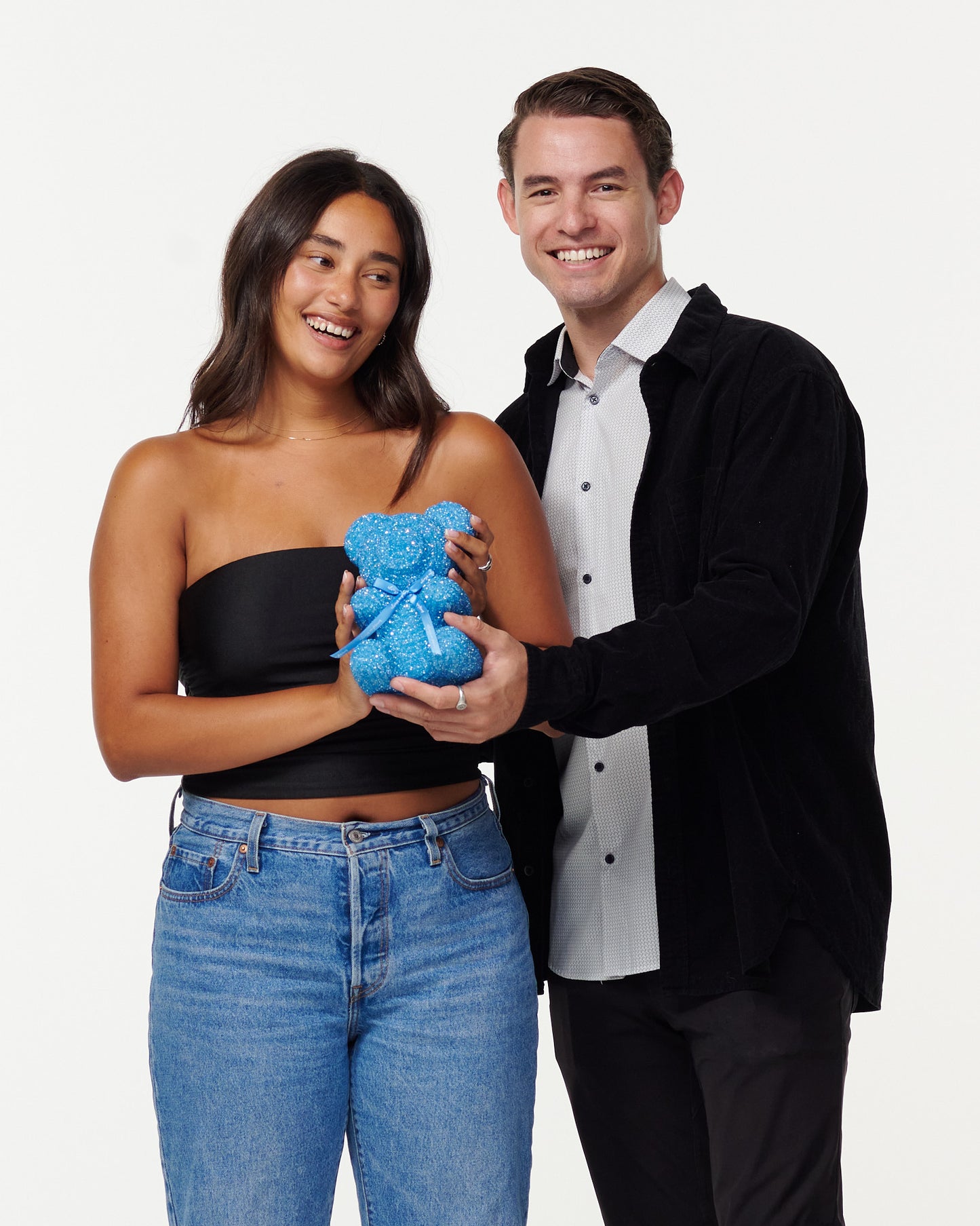 A woman in a black cropped top and blue jeans cradles a blue, sparkly teddy bear with a gentle smile. Next to her, a man in a black jacket and white shirt shares a warm smile, his hands on the teddy bear, as they both enjoy the moment together.