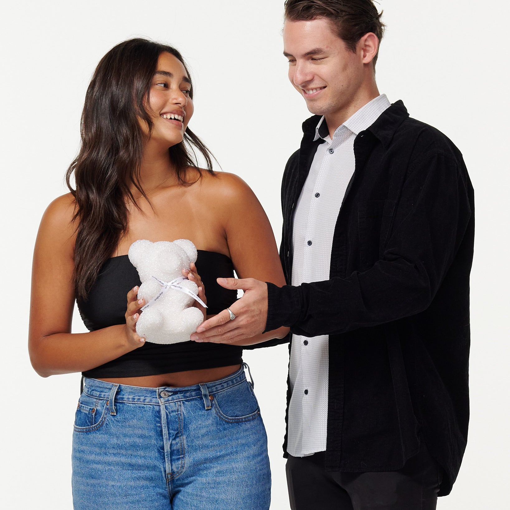 A woman in a black top and blue jeans smiles as she holds a white glitter bear with a white ribbon, while a man in a black jacket and white shirt looks on fondly.