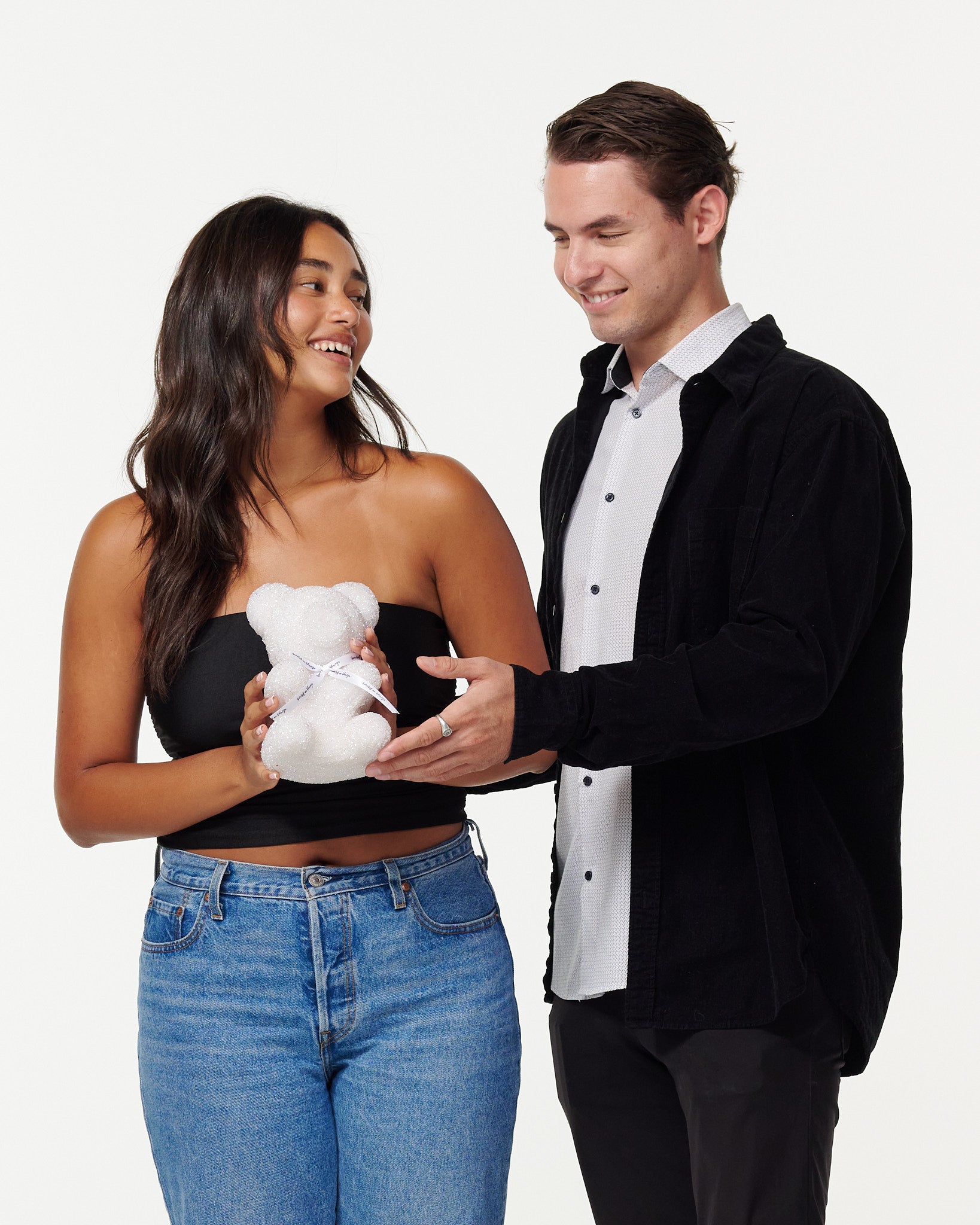 A woman in a black top and blue jeans smiles as she holds a white glitter bear with a white ribbon, while a man in a black jacket and white shirt looks on fondly.