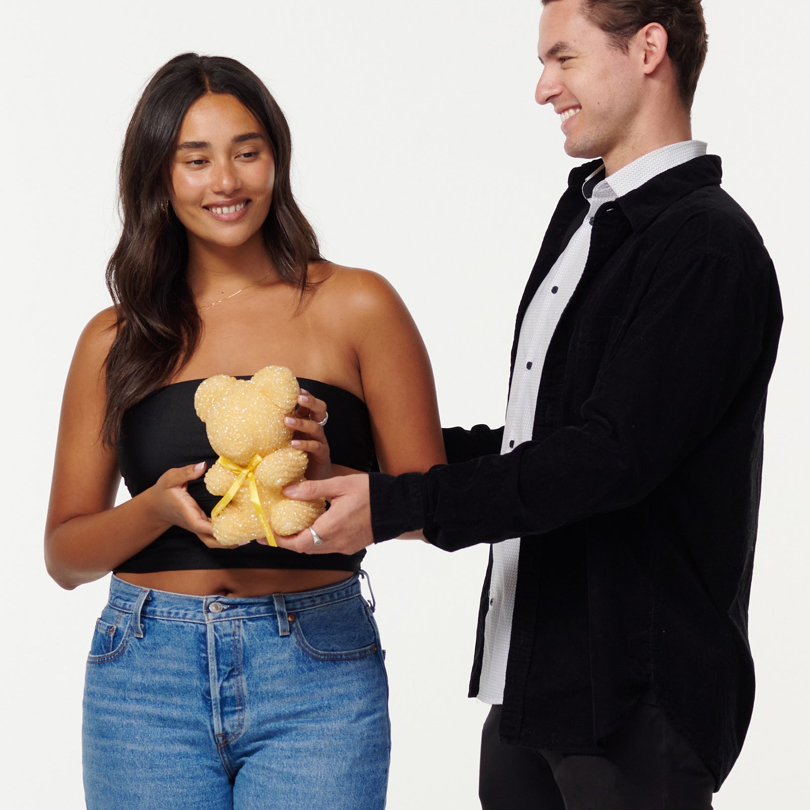 A woman and man share a moment, with the woman holding a gold-glitter bear and the man in a black jacket smiling at her. They both wear casual clothing and exude a friendly, happy demeanor.