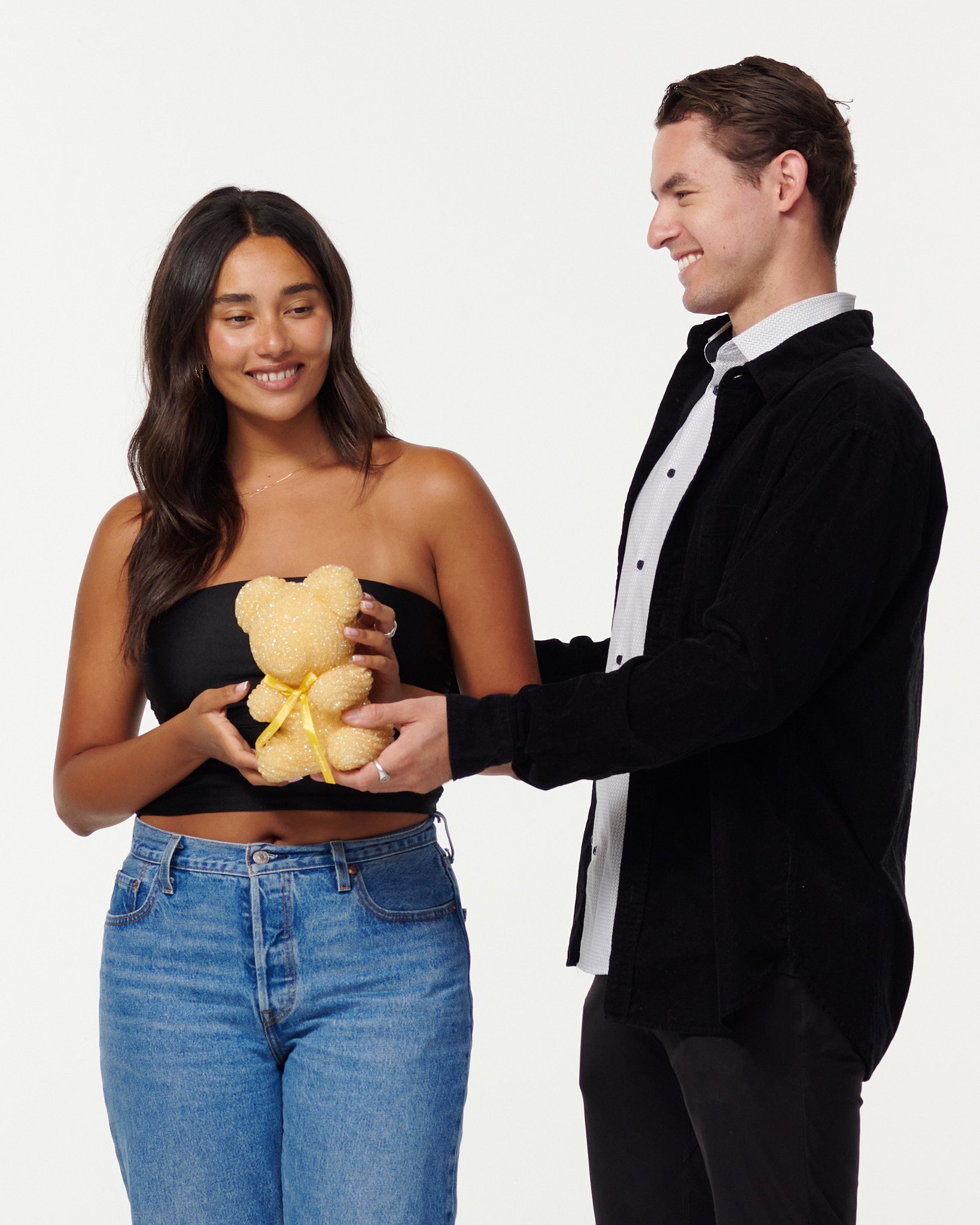 A woman and man share a moment, with the woman holding a gold-glitter bear and the man in a black jacket smiling at her. They both wear casual clothing and exude a friendly, happy demeanor.