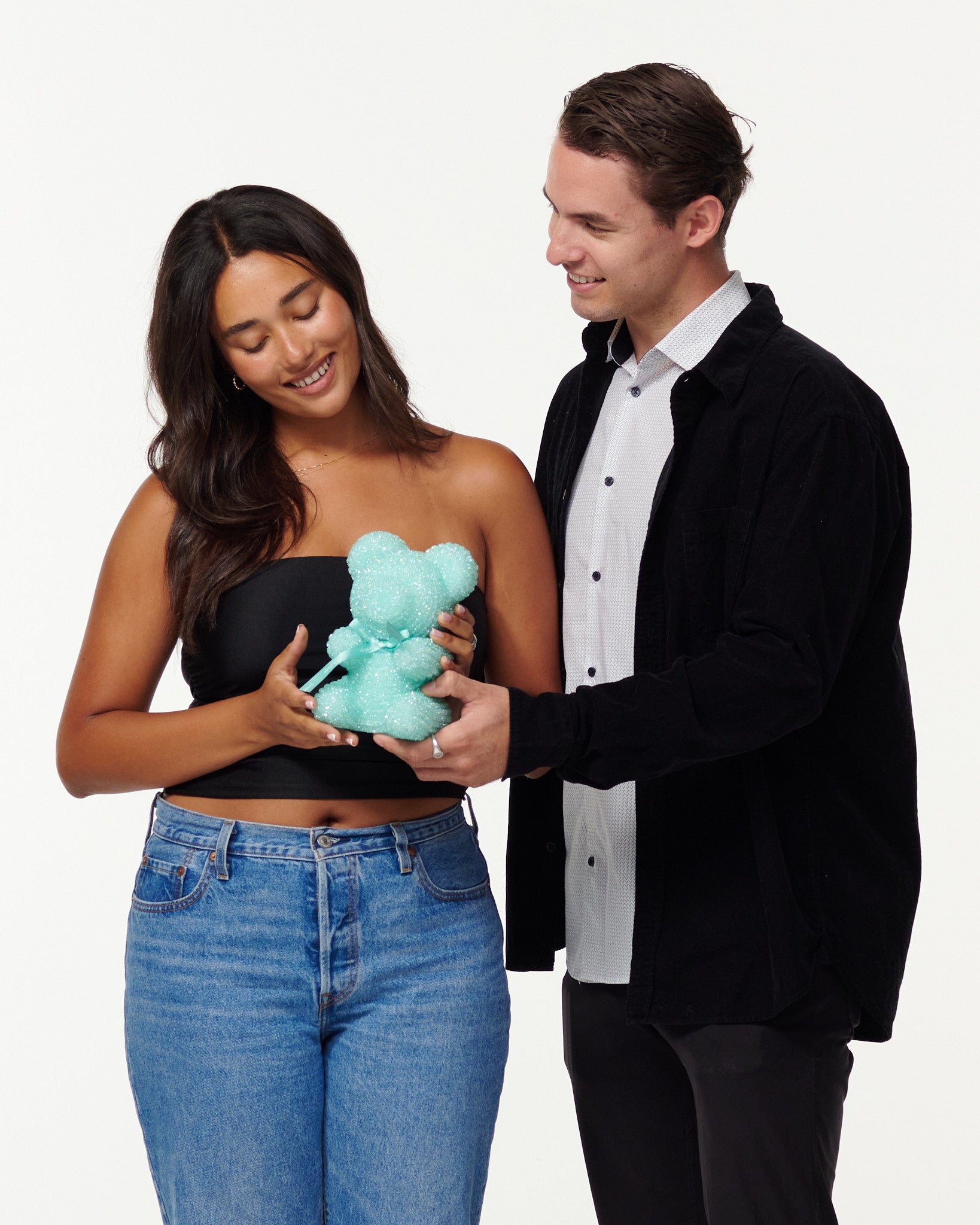 A woman in a black top and jeans admires a teal glitter bear, while a smiling man in a black jacket and white shirt gently touches the bear's arm. They appear to be in a light-hearted, affectionate interaction.