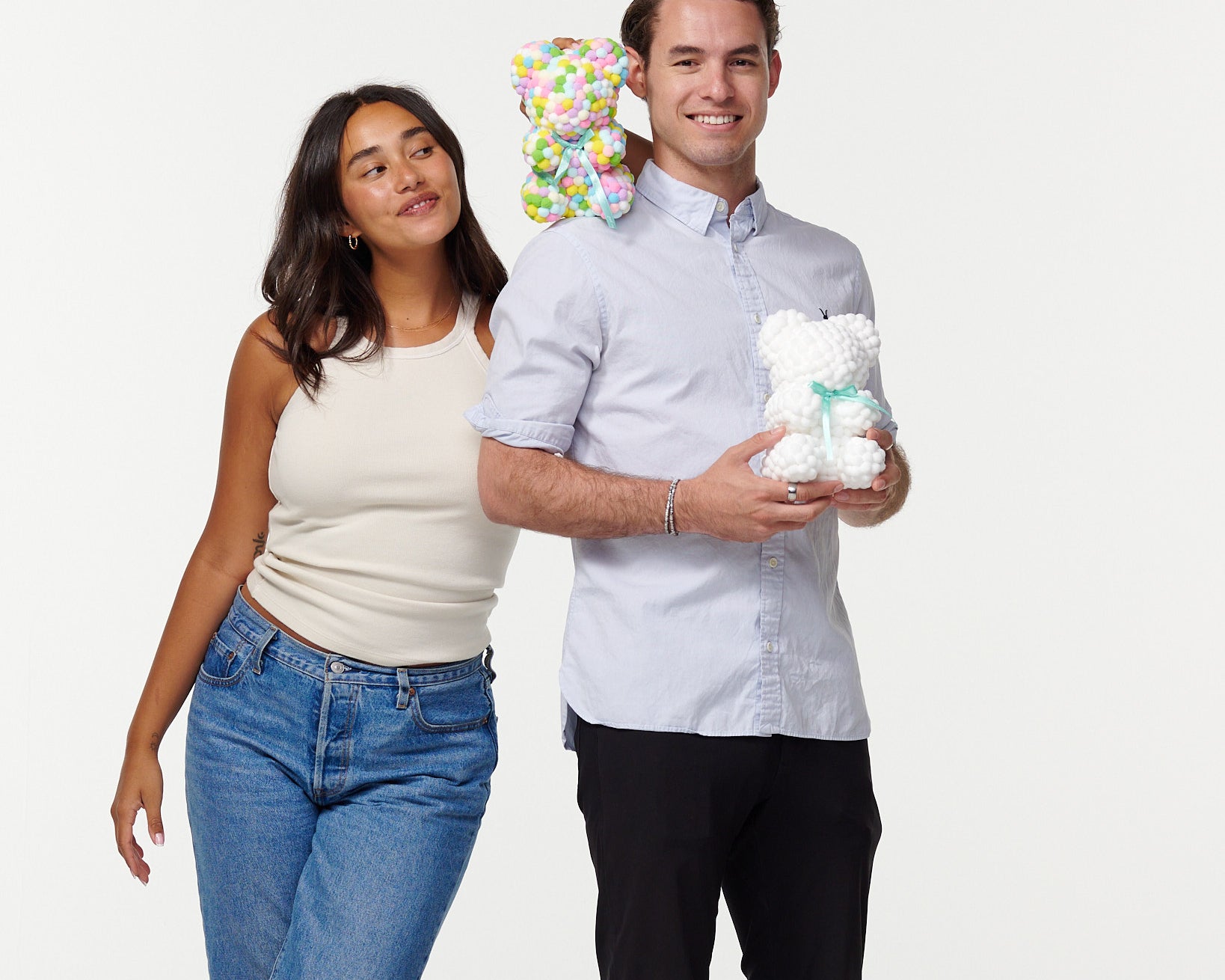 A man and woman stand together smiling; the man holds a white flower bear, and the woman rests her head on his shoulder, holding a colorful flower bear behind his head. They're casually dressed, the man in a buttoned shirt and slacks, and the woman in a tank top and jeans.