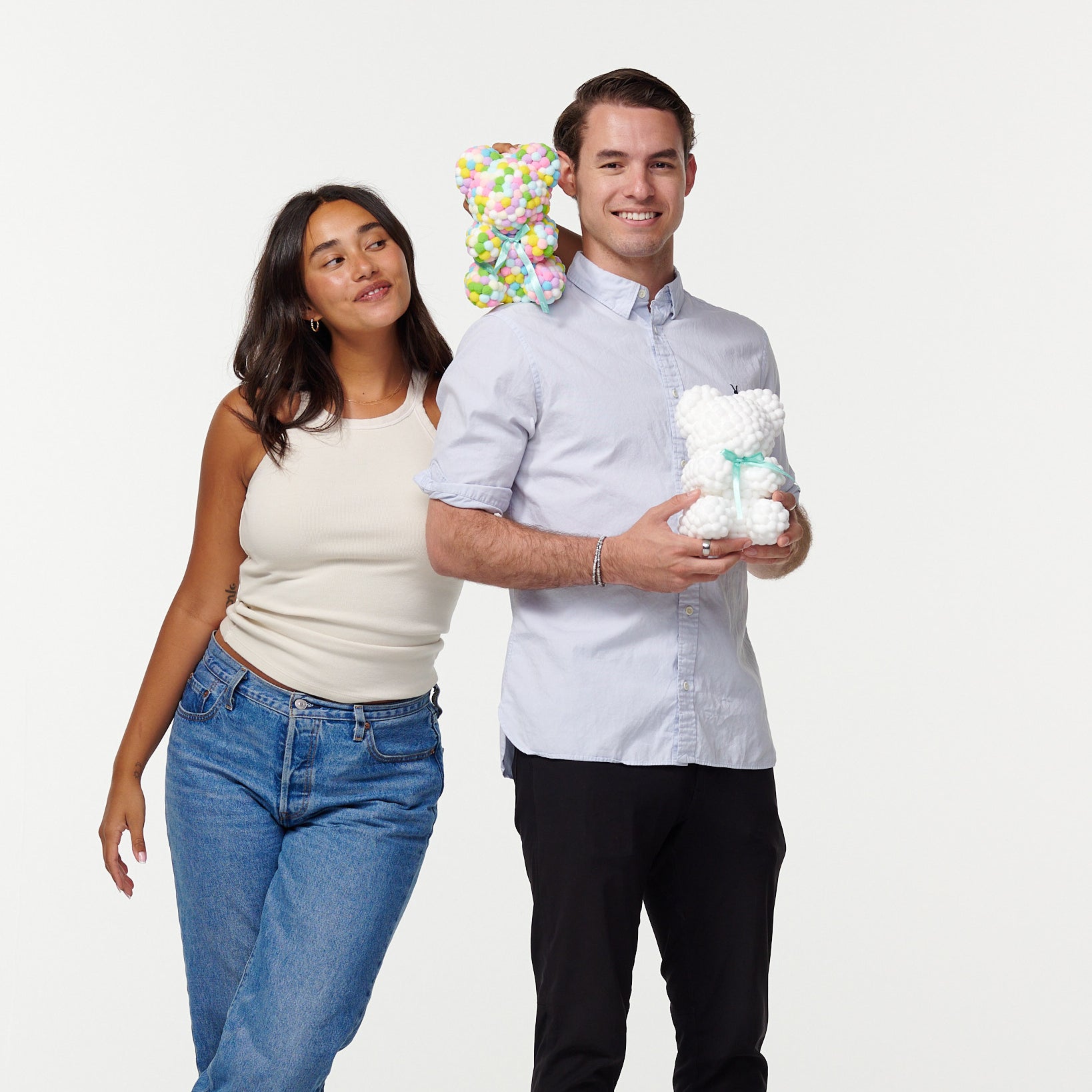 A man and woman stand together smiling; the man holds a white flower bear, and the woman rests her head on his shoulder, holding a colorful flower bear behind his head. They're casually dressed, the man in a buttoned shirt and slacks, and the woman in a tank top and jeans.