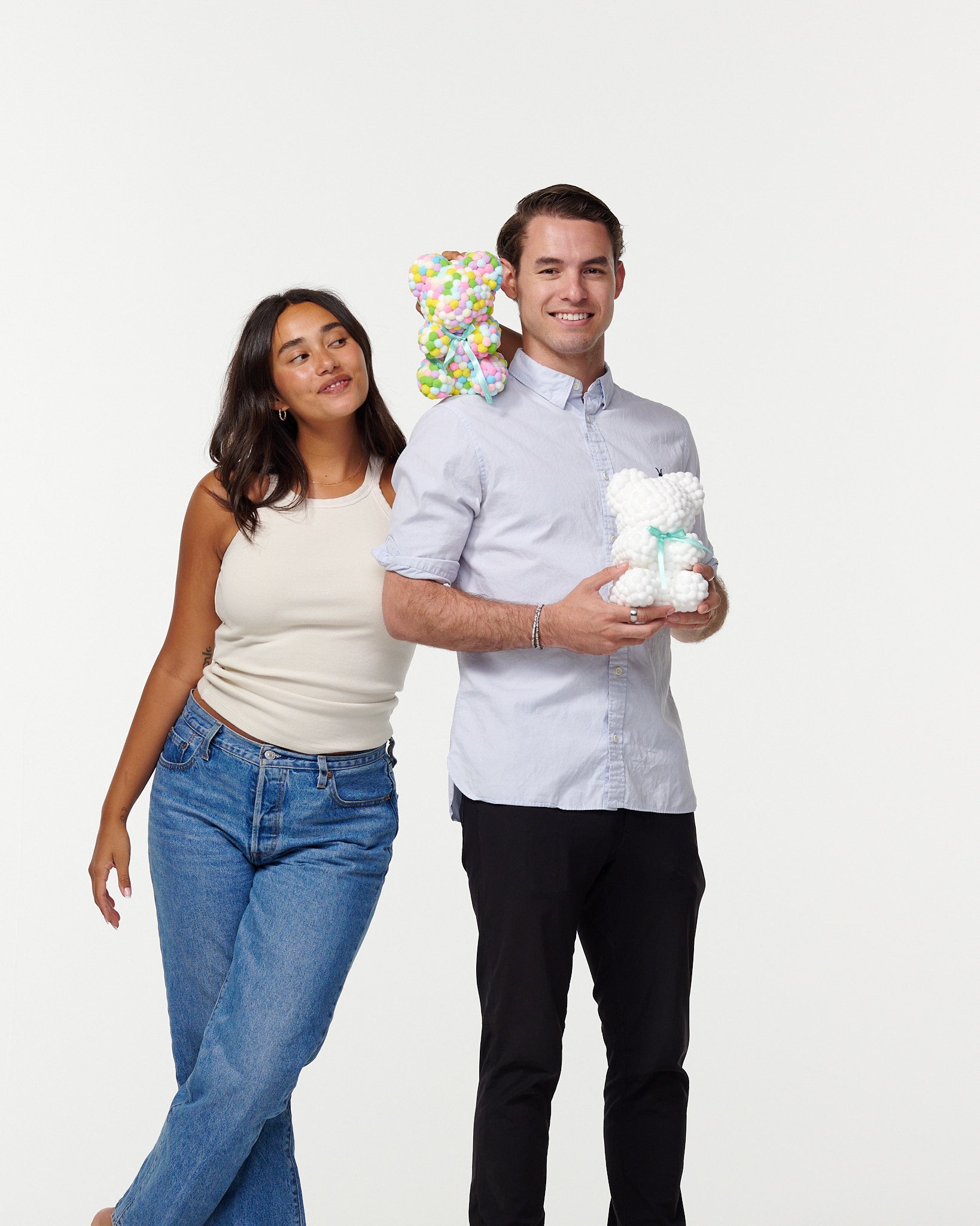 A man and woman stand together smiling; the man holds a white flower bear, and the woman rests her head on his shoulder, holding a colorful flower bear behind his head. They're casually dressed, the man in a buttoned shirt and slacks, and the woman in a tank top and jeans.