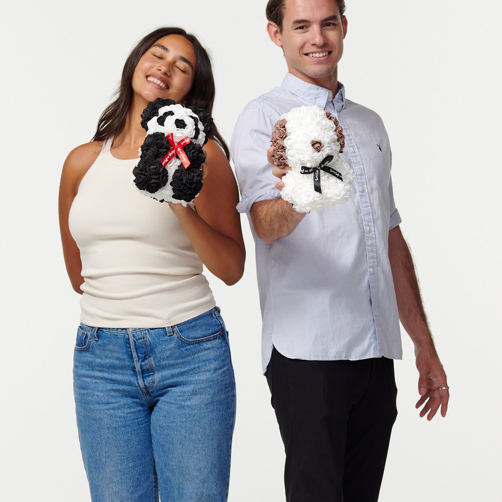 The image depicts a female model in a beige tank top and blue jeans, with her eyes closed and a content smile, holding a black and white flower panda with a red ribbon. Beside her, a male model in a light blue shirt and black pants smiles at the camera, holding a white flower bear with a black ribbon. Both models are presenting the artificial flower animals as treasured items, suggesting a joyful and affectionate context