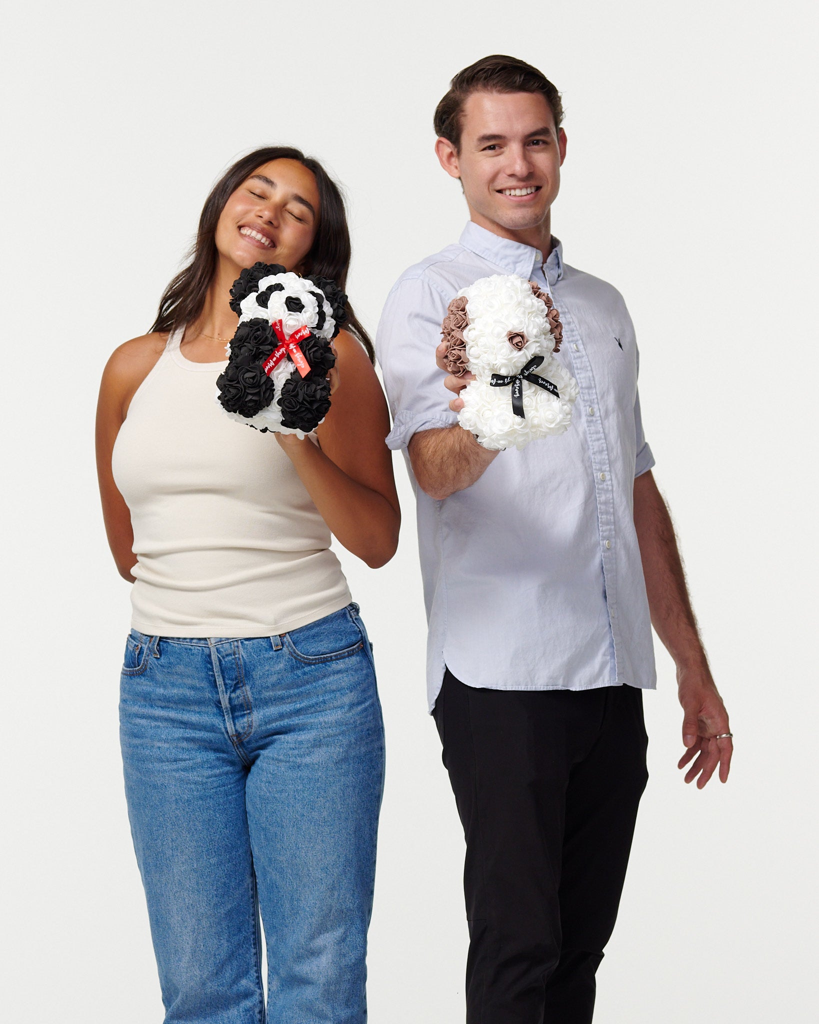 The image depicts a female model in a beige tank top and blue jeans, with her eyes closed and a content smile, holding a black and white flower panda with a red ribbon. Beside her, a male model in a light blue shirt and black pants smiles at the camera, holding a white flower bear with a black ribbon. Both models are presenting the artificial flower animals as treasured items, suggesting a joyful and affectionate context