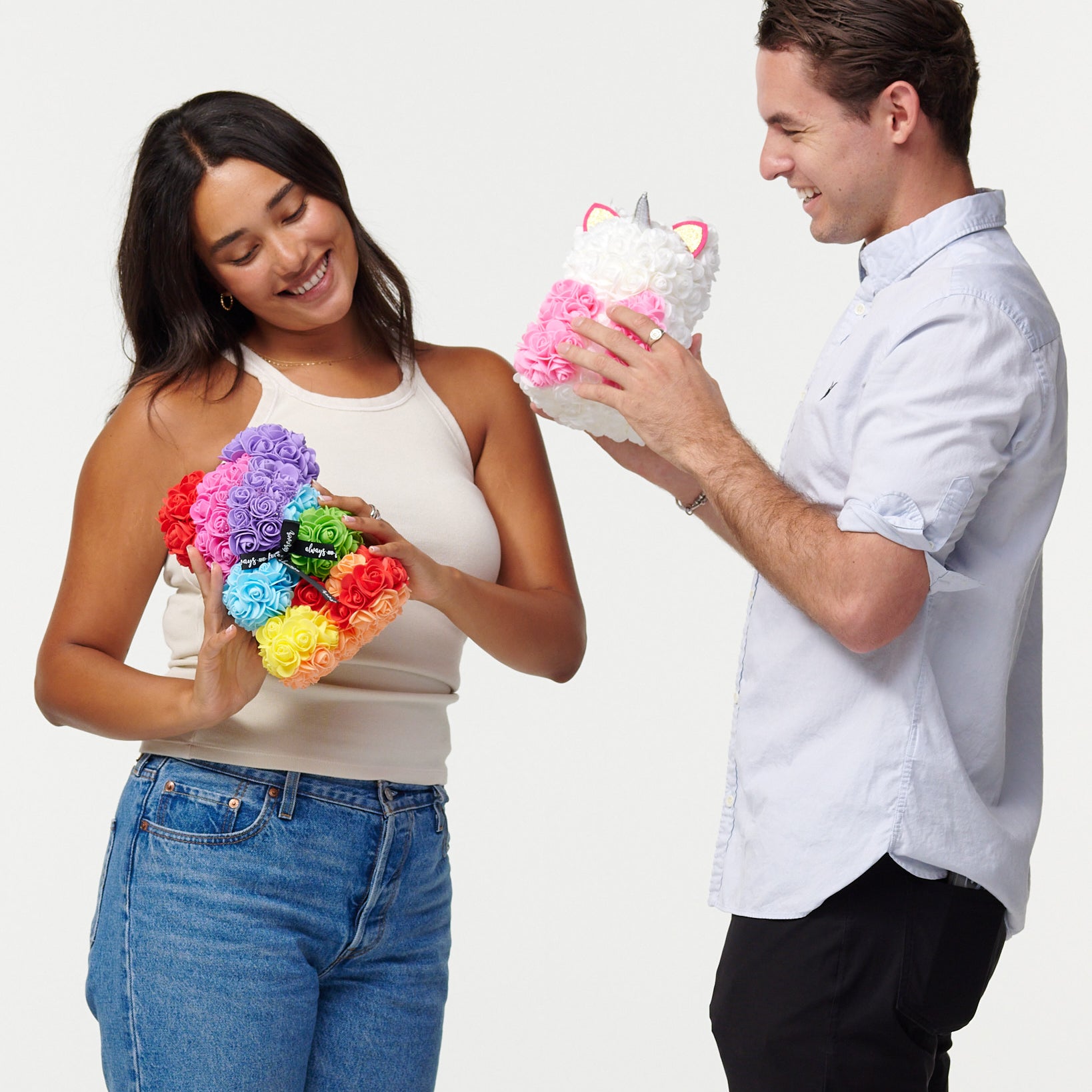 A female model in a beige tank top and blue jeans is holding a multicolored flower bear, looking down at it with a smile. Next to her, a male model in a light blue button-up shirt and black pants is admiring a white flower unicorn he holds, which has pink flowers for its mane and a silver horn. They both appear cheerful and are engaging with the artificial flower animals, creating a light-hearted and happy scene.