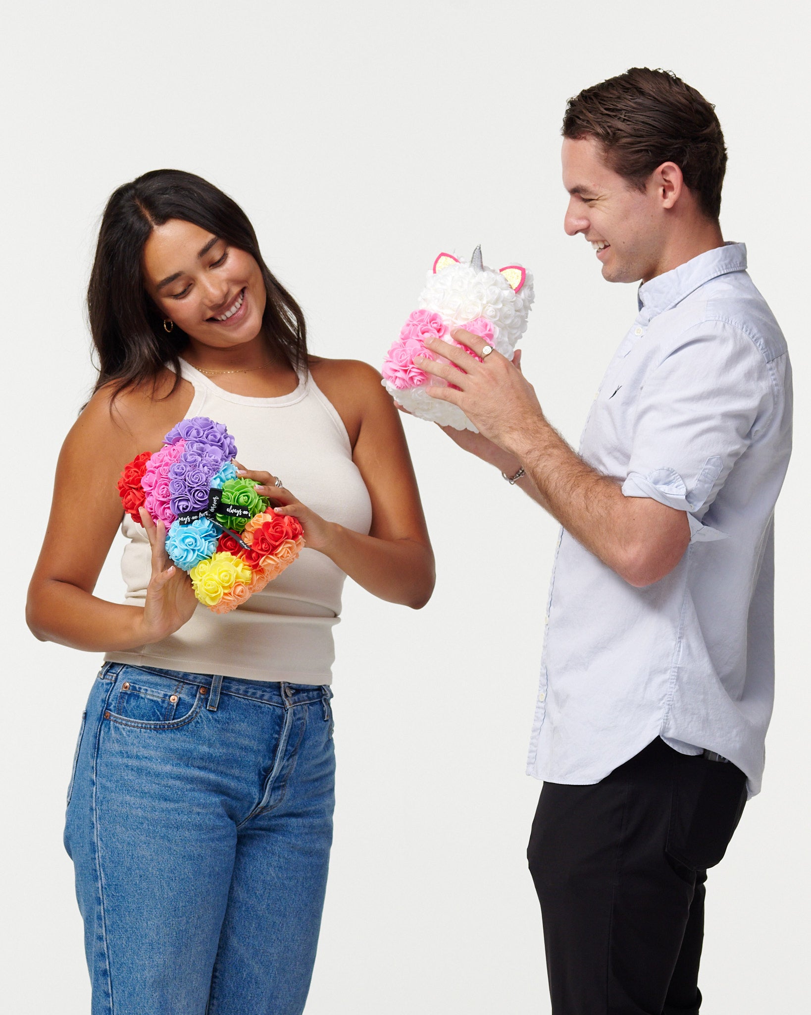 A female model in a beige tank top and blue jeans is holding a multicolored flower bear, looking down at it with a smile. Next to her, a male model in a light blue button-up shirt and black pants is admiring a white flower unicorn he holds, which has pink flowers for its mane and a silver horn. They both appear cheerful and are engaging with the artificial flower animals, creating a light-hearted and happy scene.