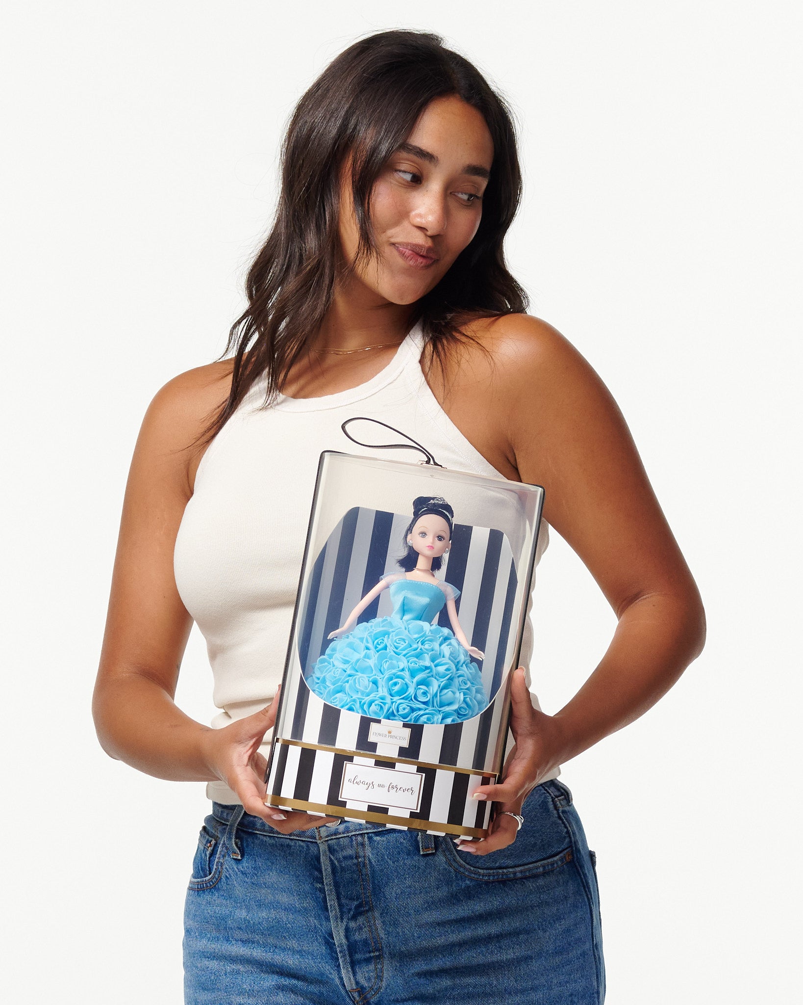 A woman is pictured holding a boxed doll. The doll within the packaging is styled with black hair, green eyes, and is dressed in a blue rose-adorned gown. The box displays black and white stripes, a gold trim, and a black ribbon with the text "forever always" on the top. The woman is casually dressed in a white tank top and blue jeans.