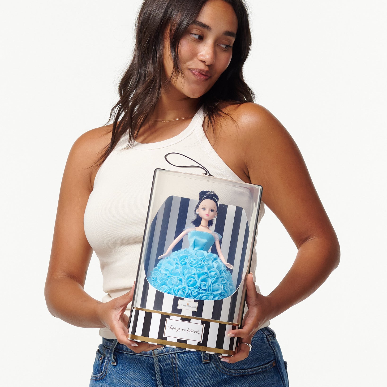 A woman is pictured holding a boxed doll. The doll within the packaging is styled with black hair, green eyes, and is dressed in a blue rose-adorned gown. The box displays black and white stripes, a gold trim, and a black ribbon with the text "forever always" on the top. The woman is casually dressed in a white tank top and blue jeans.